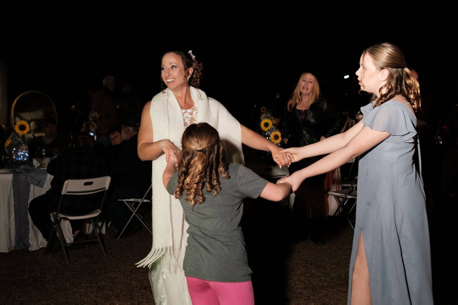 Bride in white wrap dancing joyfully with young children at an outdoor evening wedding reception.