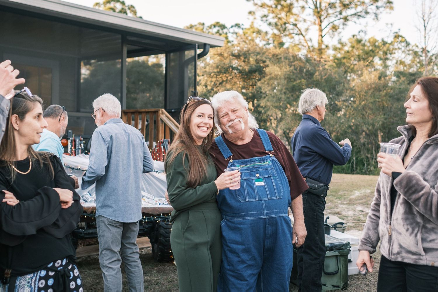 Young woman hugs older man in overalls at outdoor wedding reception near rustic cabin in woods.