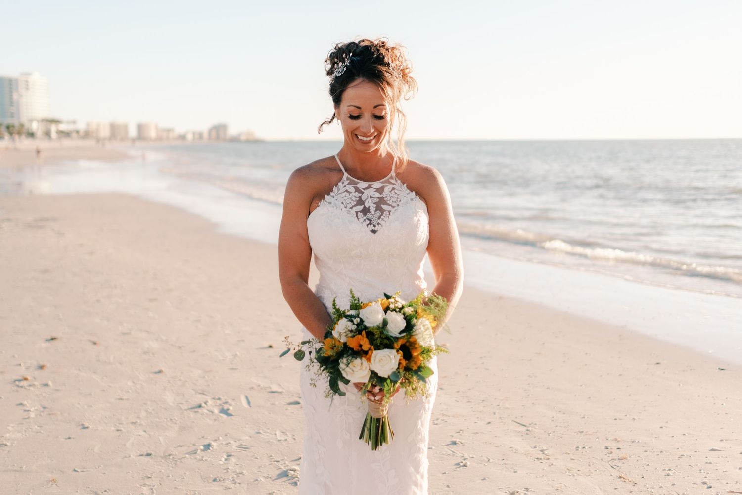 Bride in halter lace gown holding white and yellow bouquet smiling on sandy beach at sunset.