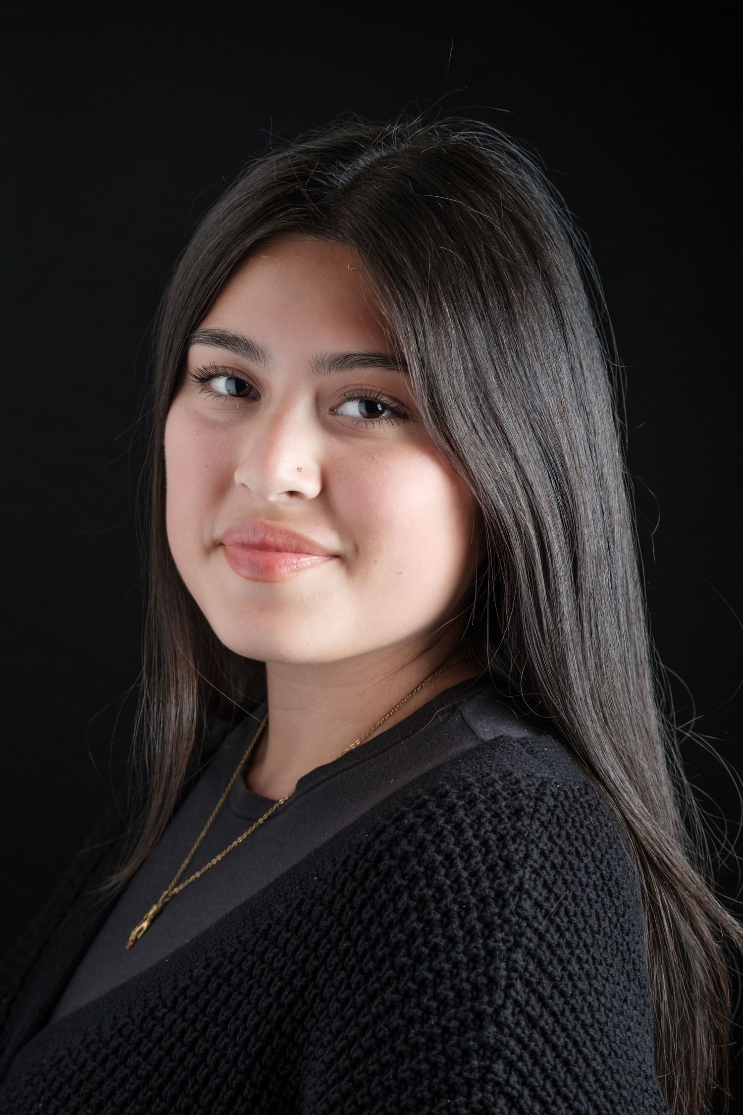 Young woman with long dark hair smiling, wearing a black top and gold necklace, against a dark background.