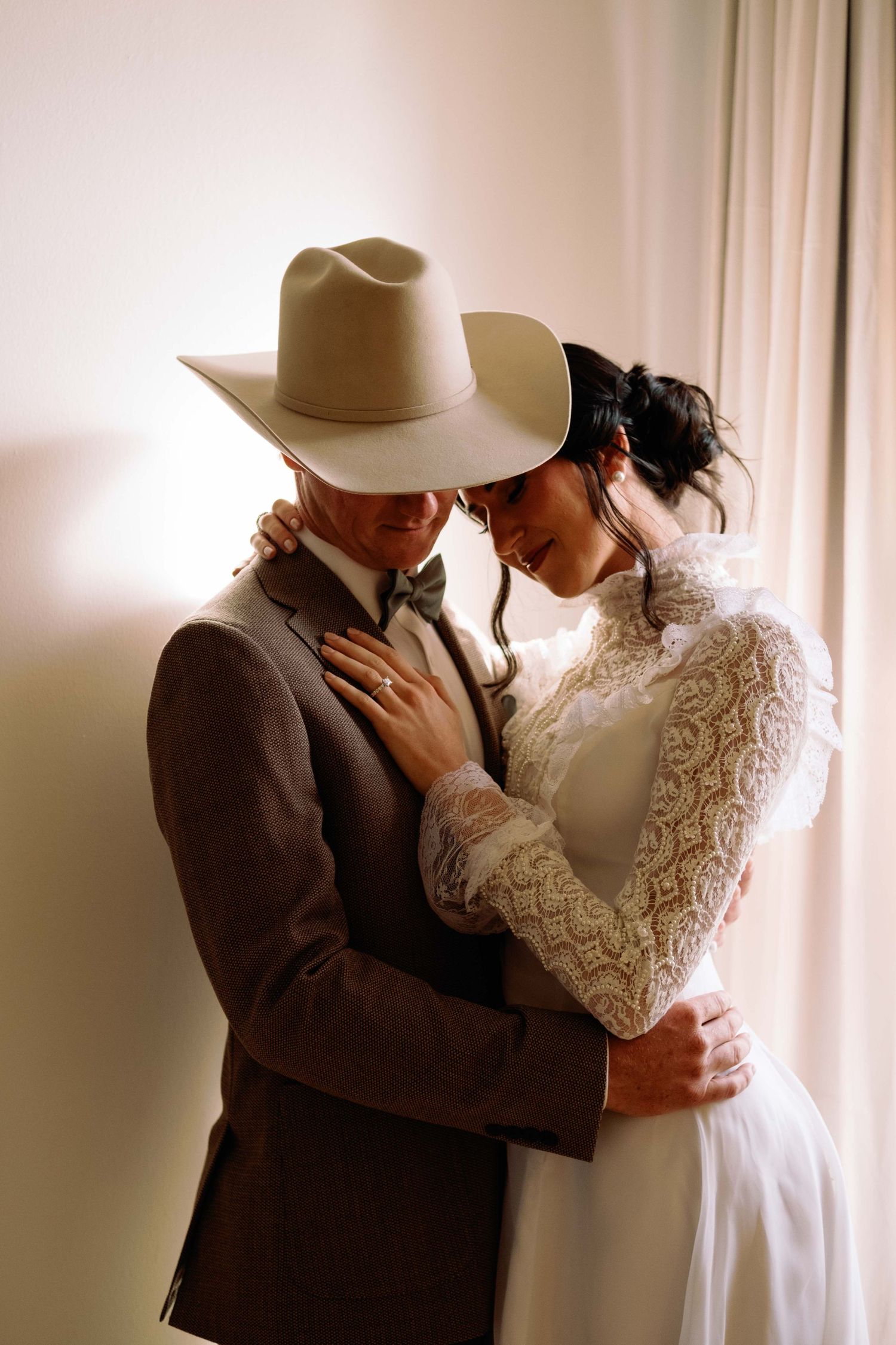 A bride in lace dress and groom in brown suit with cowboy hat share an intimate embrace near a sunlit window.