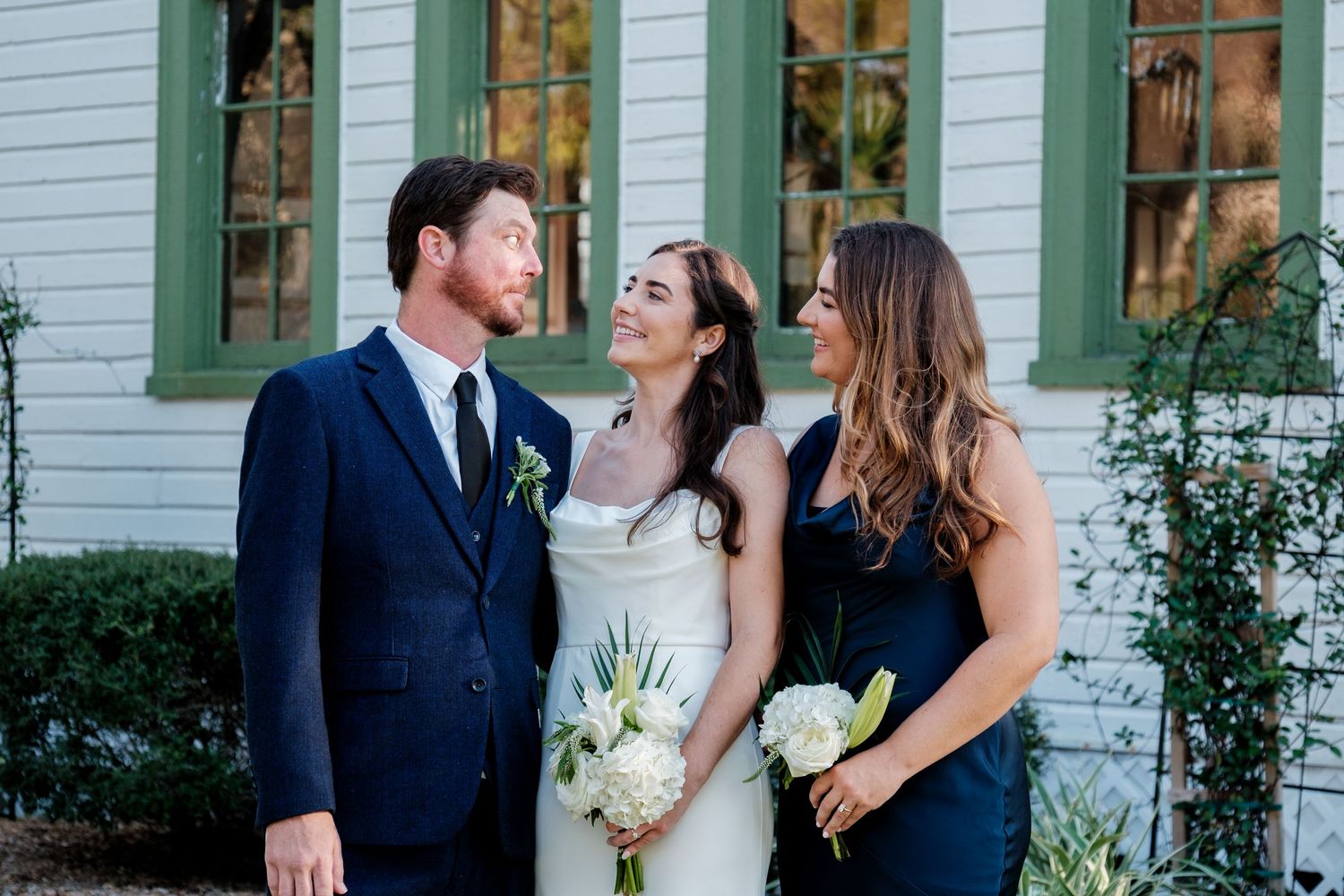 Wedding party members share a joyful moment together outside a white building with green shutters.