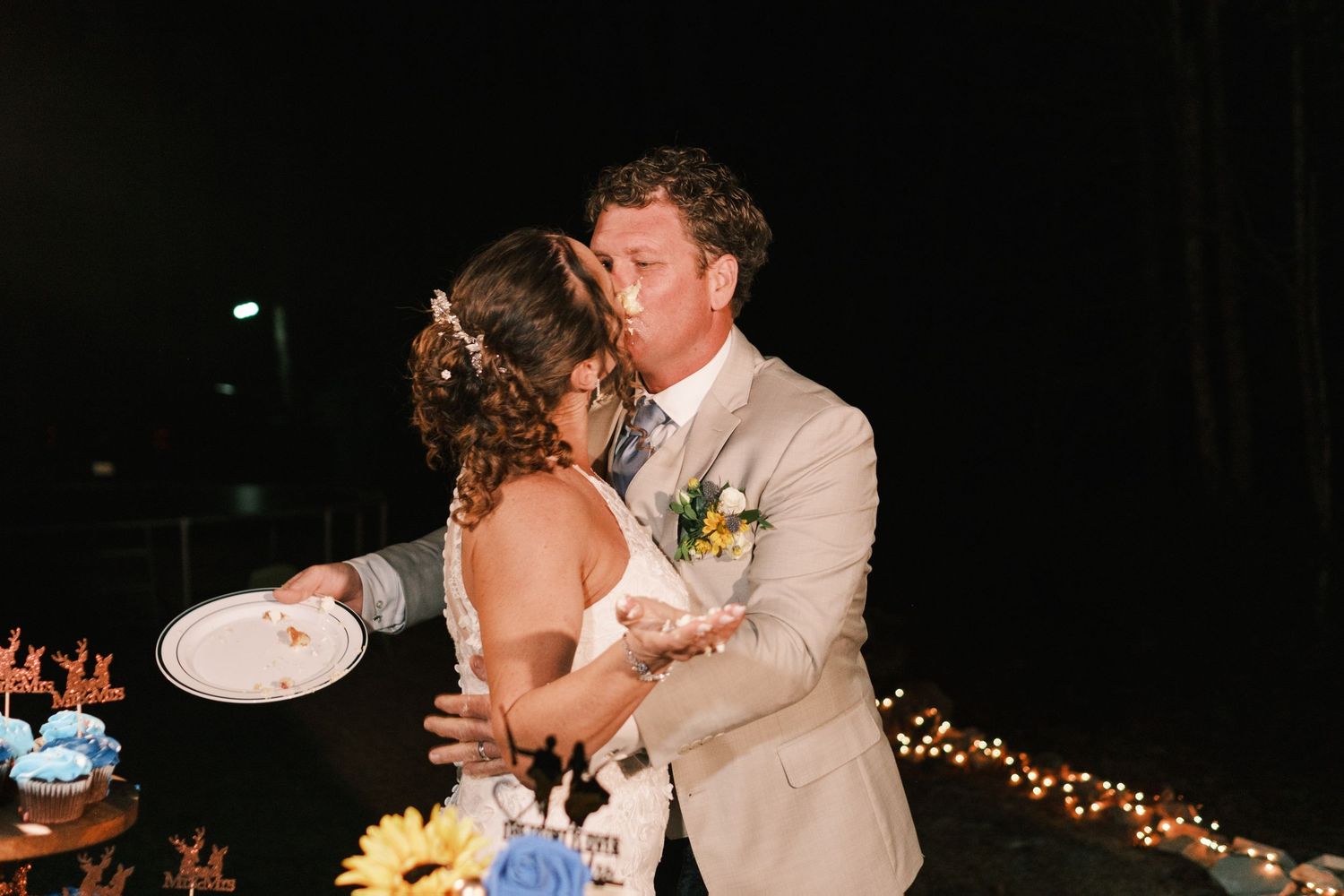 Bride and groom share a kiss while cutting their wedding cake at a romantic evening reception with string lights.
