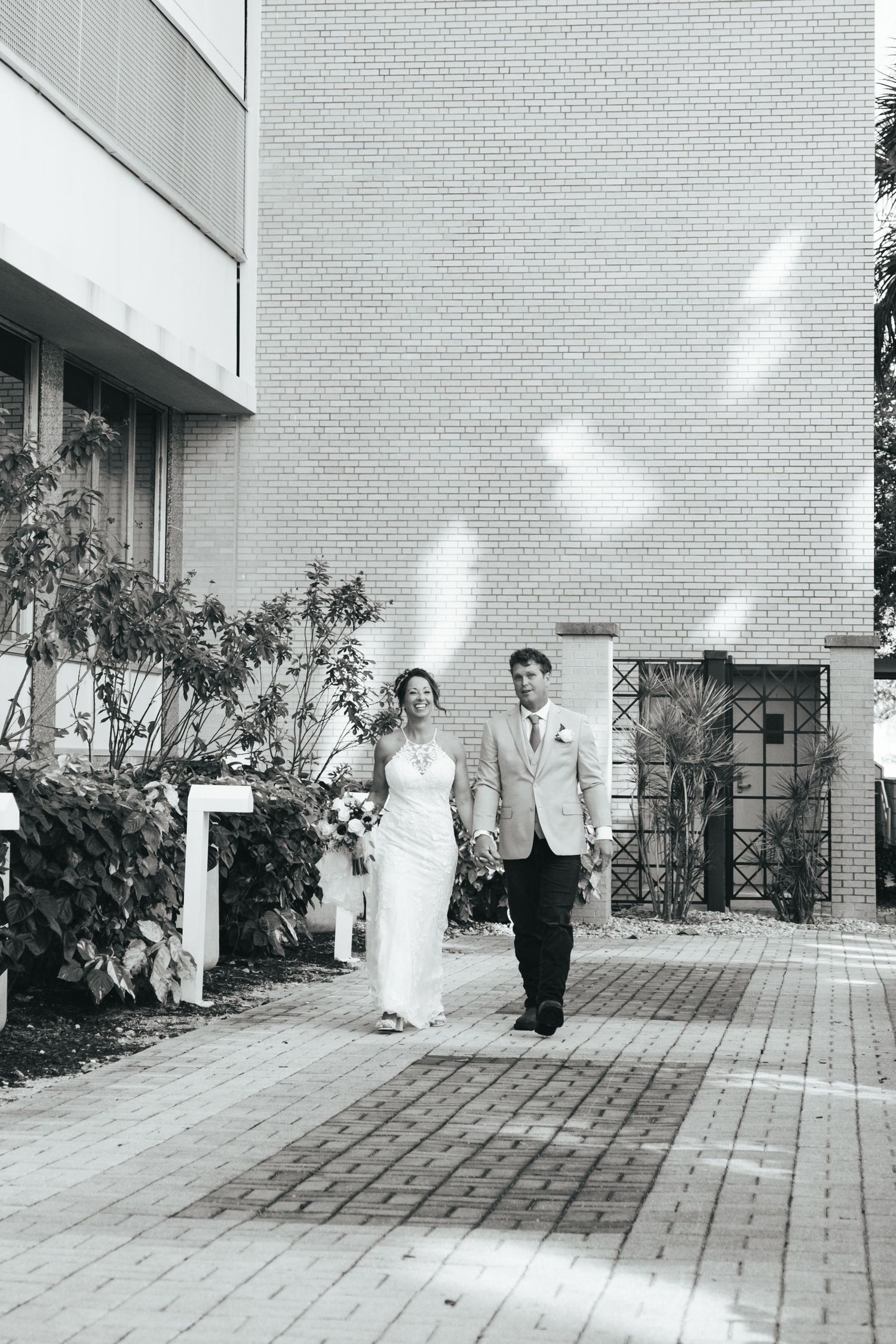 Bride and groom walking together on brick pathway with white petals falling in black and white photo.