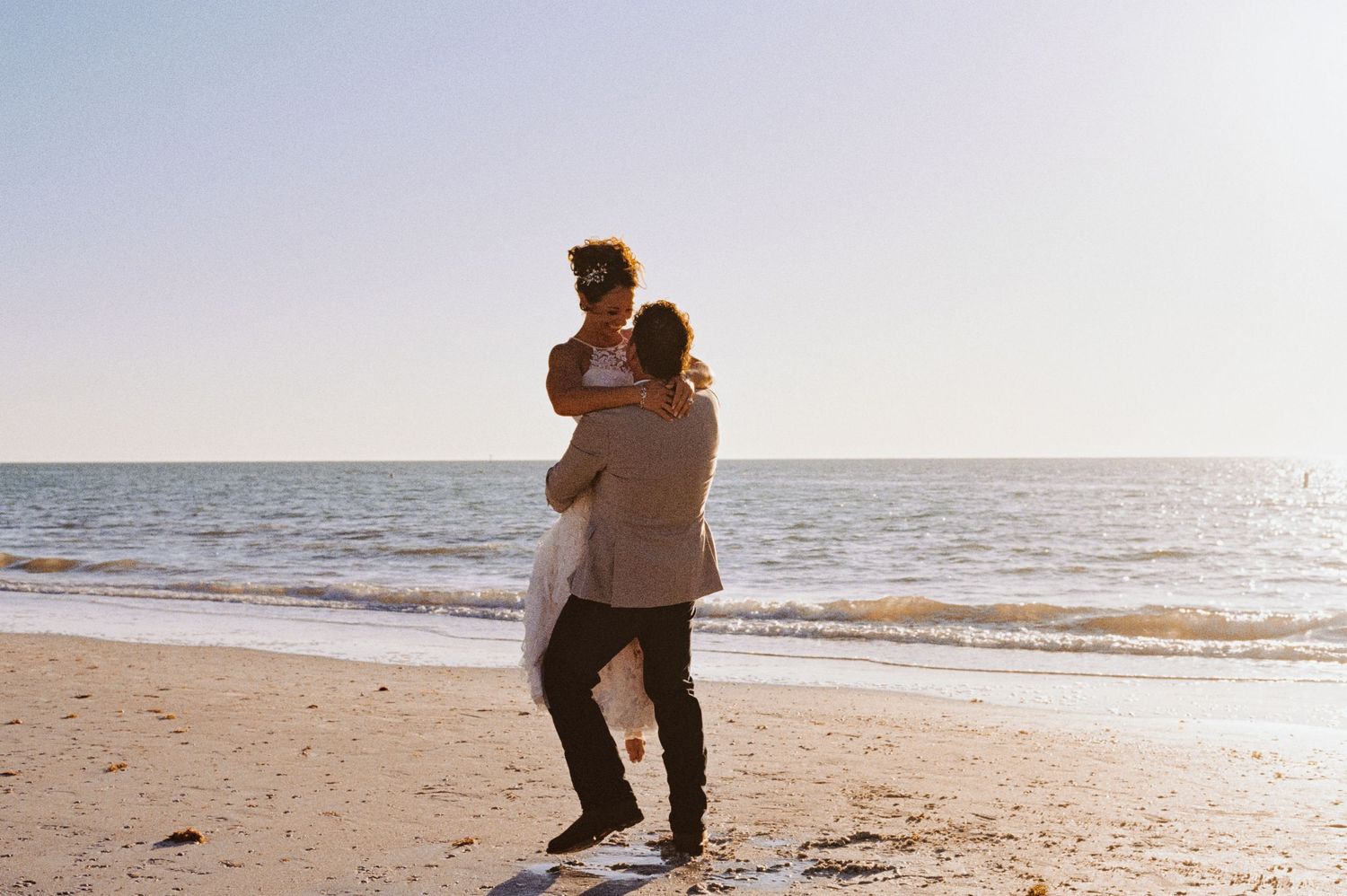 Couple embracing romantically on a sunny beach at sunset with ocean waves in the background.