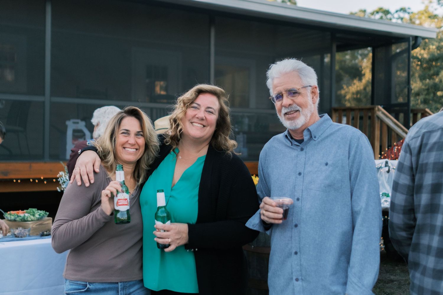 Three smiling wedding guests pose together holding drinks at an outdoor evening reception.