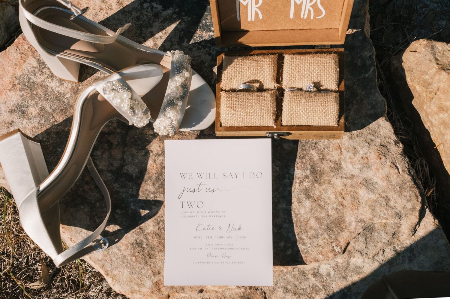 Wedding flat lay featuring invitation, ring boxes, ribbon, and Mr and Mrs wooden box on stone.
