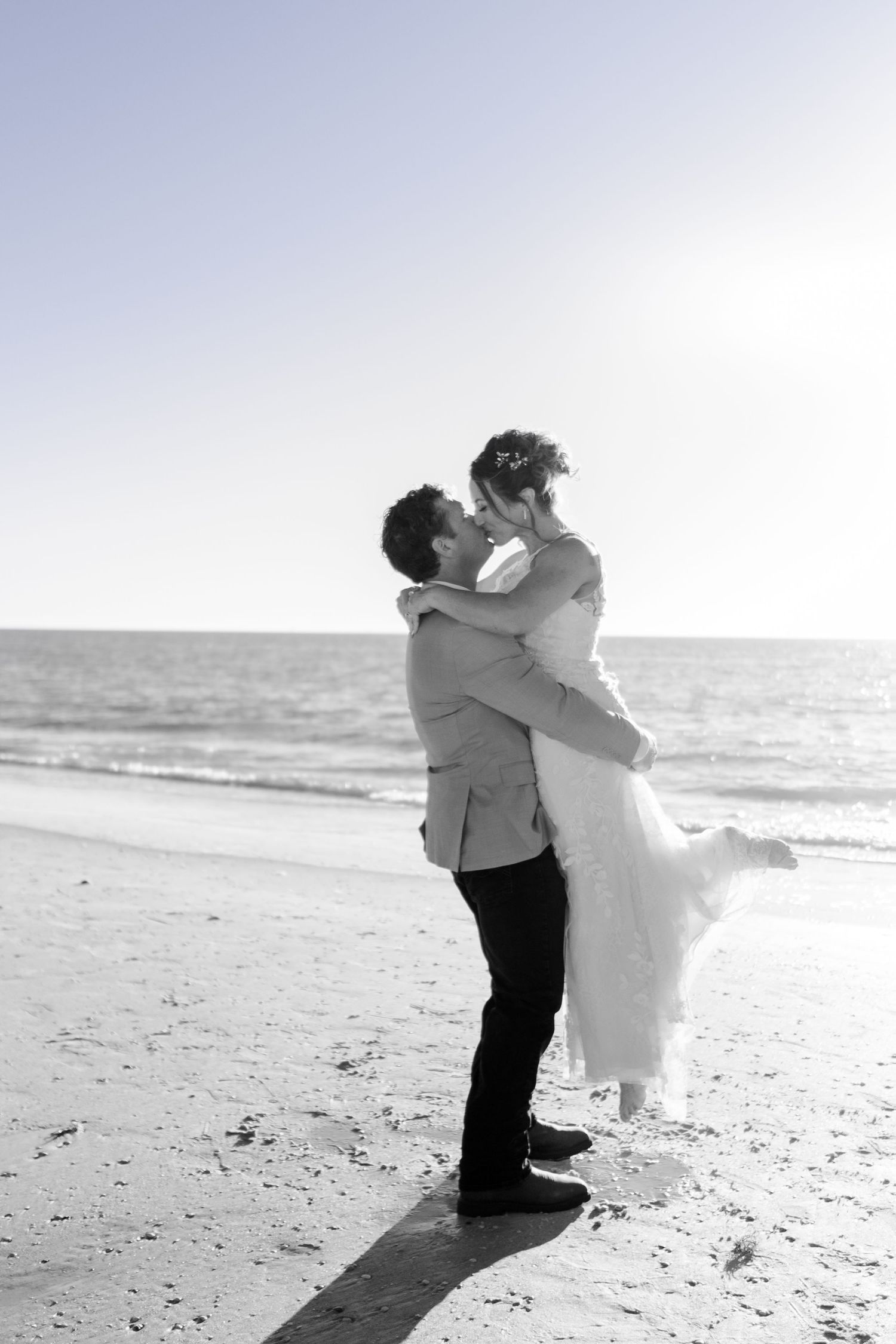 Black and white photo of groom lifting bride for a kiss on the beach.