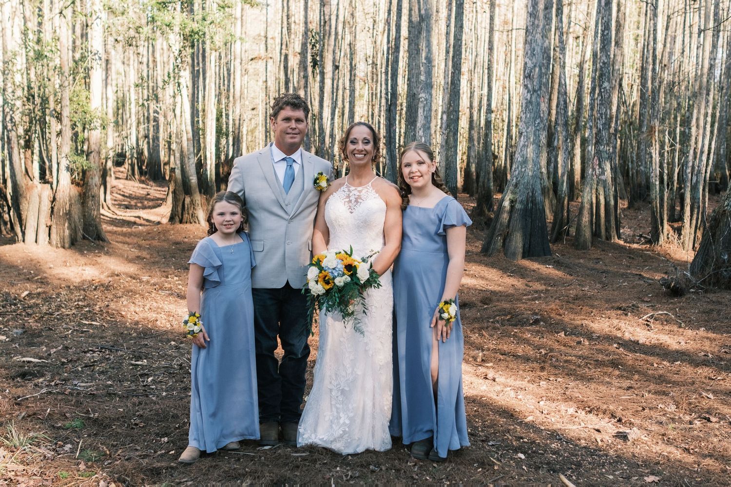 Wedding party poses in a forest with bride in white gown, groom in tan suit, and bridesmaids in dusty blue dresses.