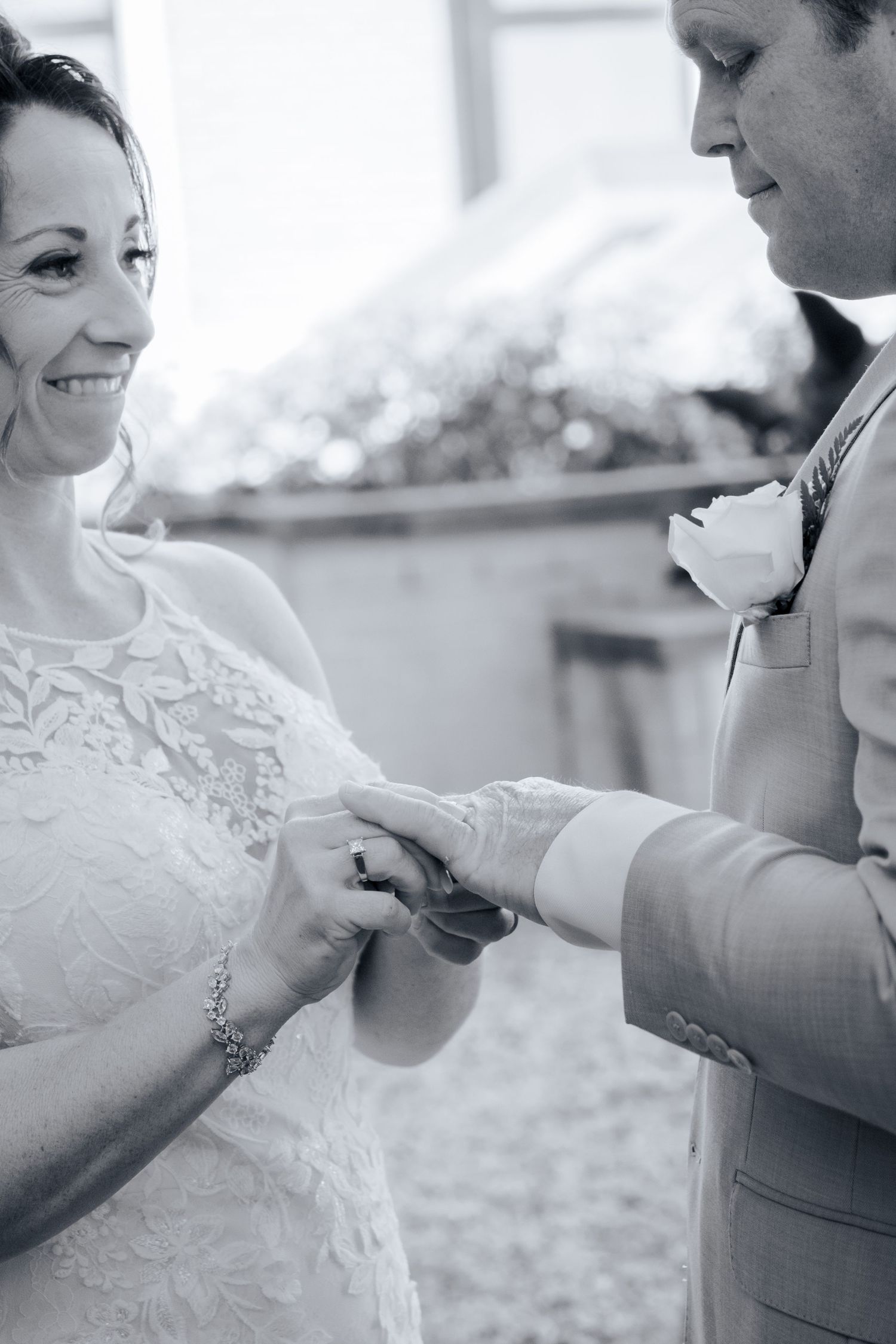 Bride smiling as groom places wedding ring on her finger during ceremony in black and white photo.
