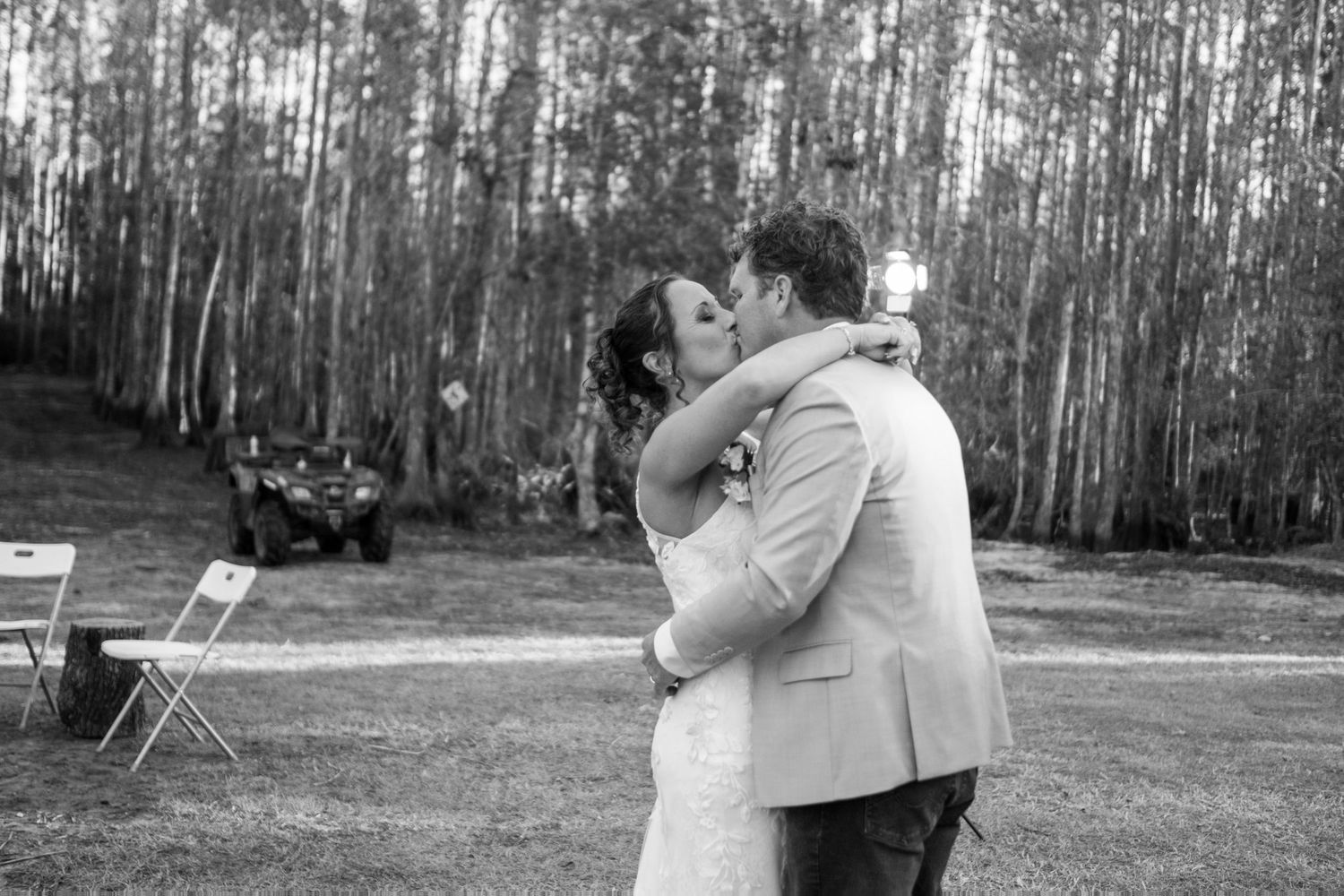 Black and white photo of bride and groom sharing first kiss outdoors surrounded by tall trees at wedding.