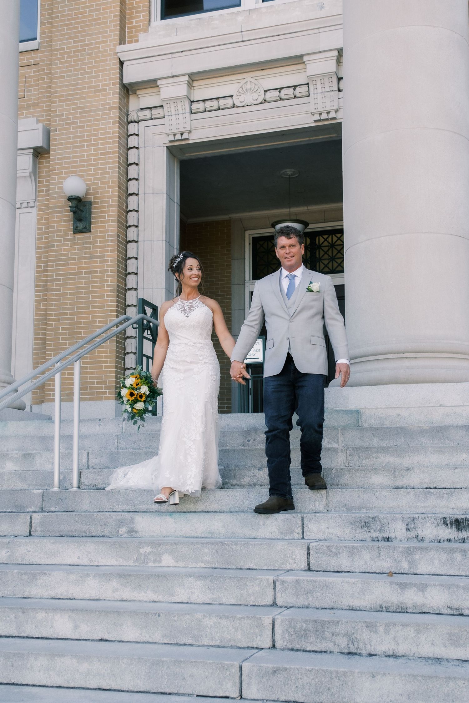Bride and groom walk hand in hand down steps of historic building after ceremony.