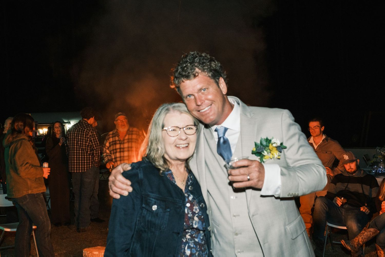 Groom in tan suit poses with older woman in front of bonfire at wedding reception.
