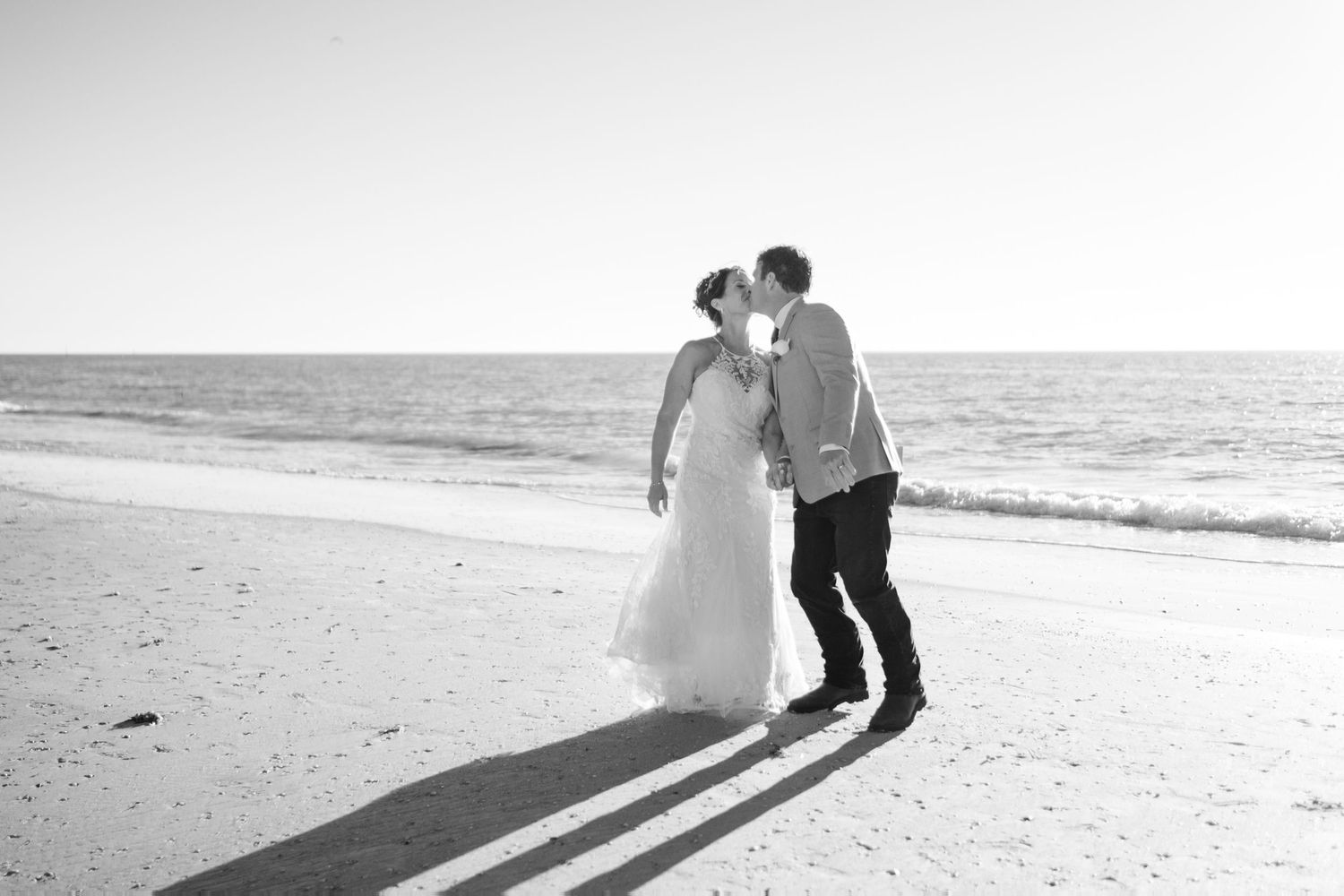 Black and white photo of bride and groom sharing a kiss on a sandy beach with ocean waves behind them.