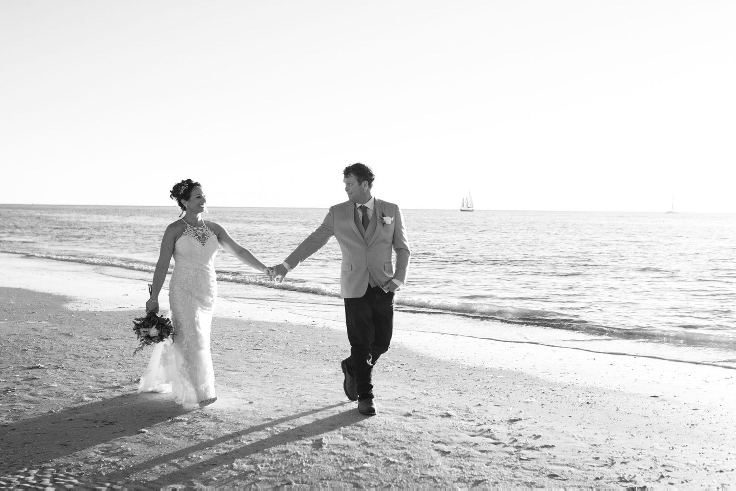 Black and white photo of newlyweds walking hand in hand along a sunny beach.