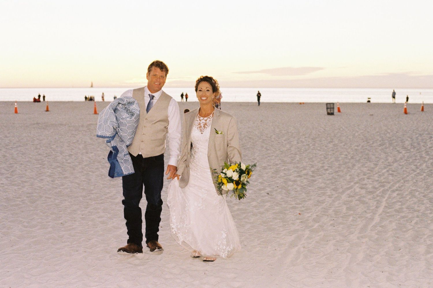 Bride and groom walk hand in hand on a wide sandy beach at sunset, bride holding yellow bouquet.