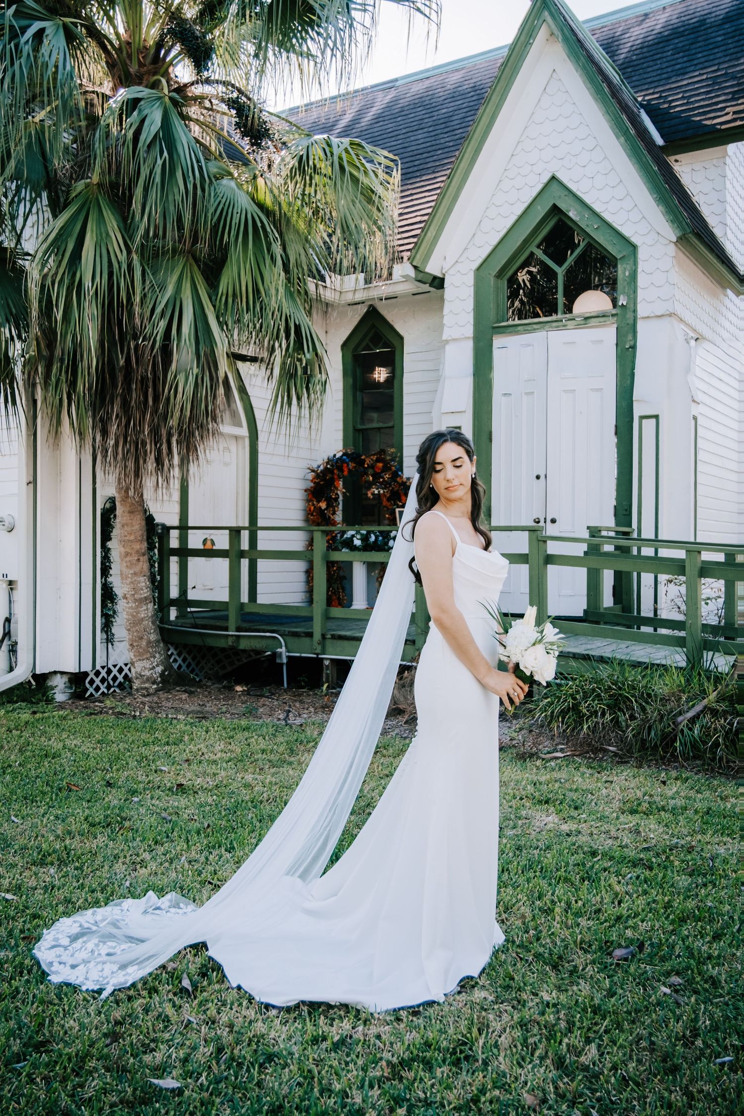A bride in a flowing white dress poses in front of a white church building with green trim and palm trees.