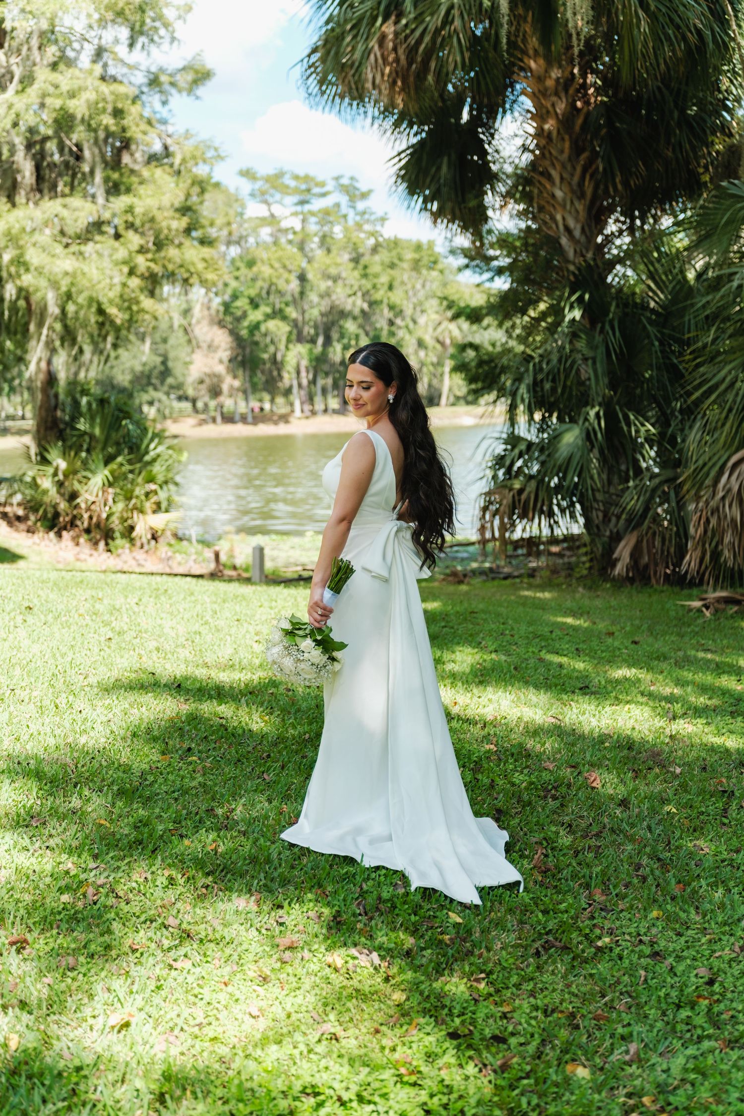 Bride in flowing white dress poses in lush garden setting near a lake.