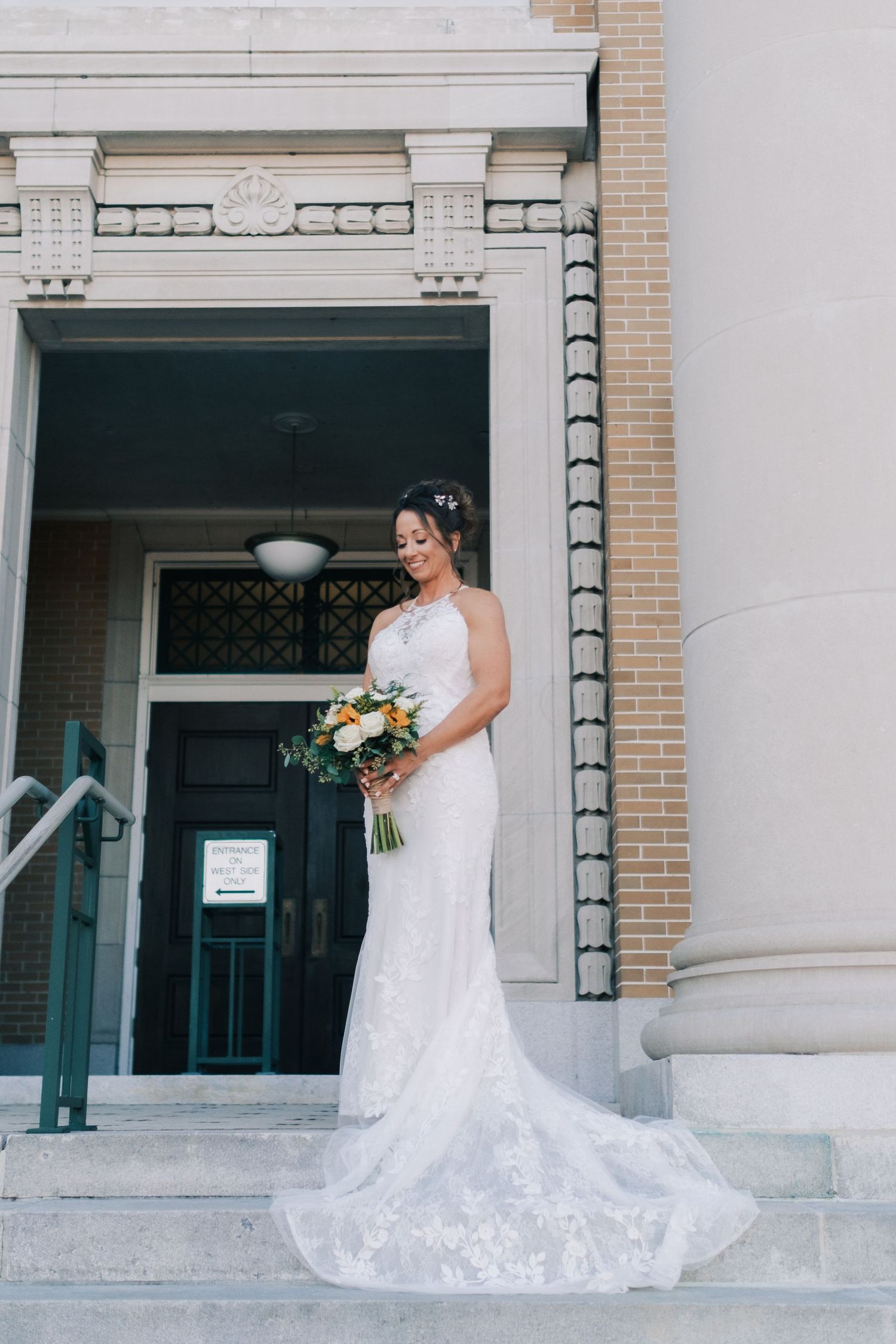 Elegant bride in lace wedding dress looking down at bouquet on building steps.