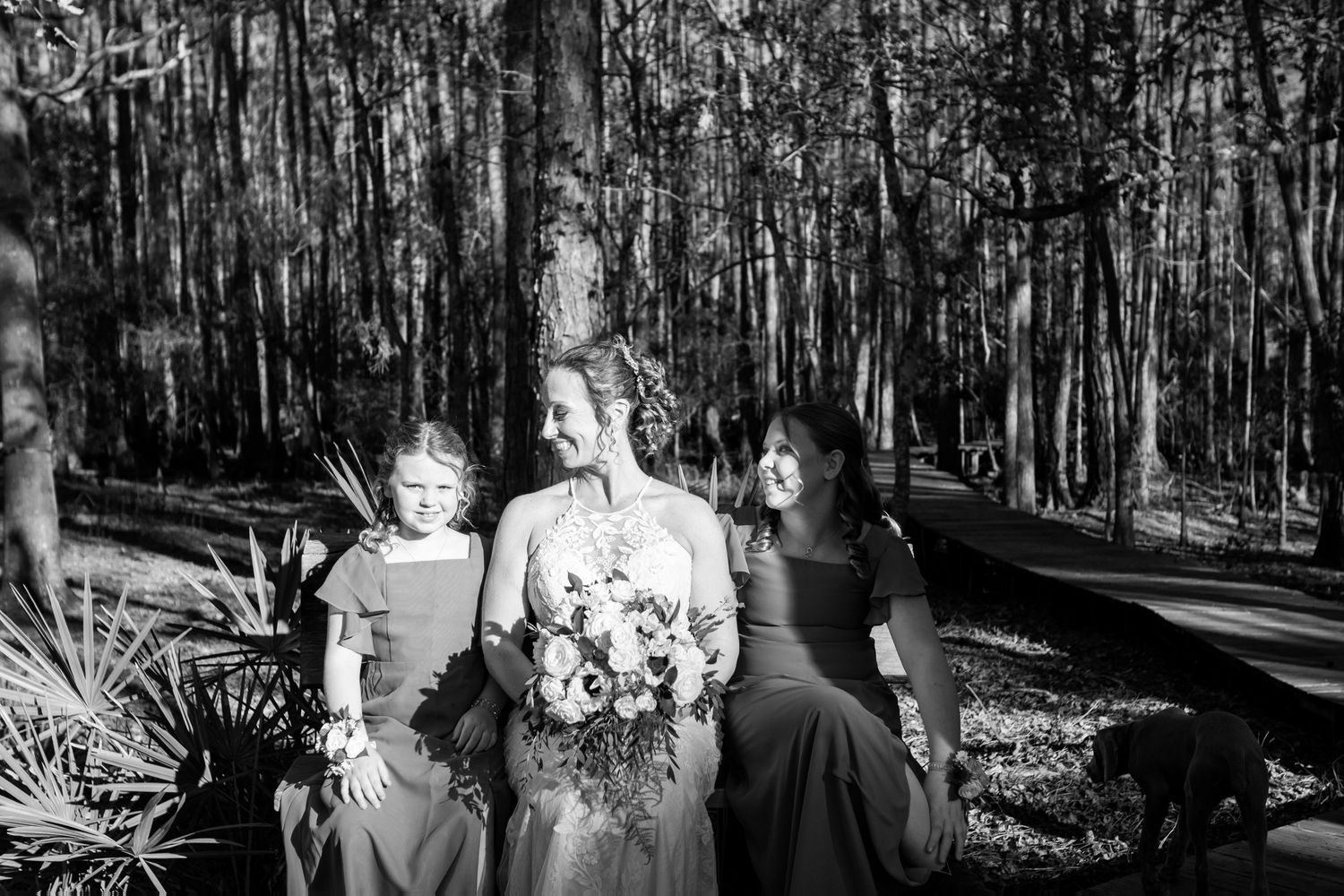 Black and white photo of a bride and two bridesmaids in dark gowns sharing a joyful moment in a forest.