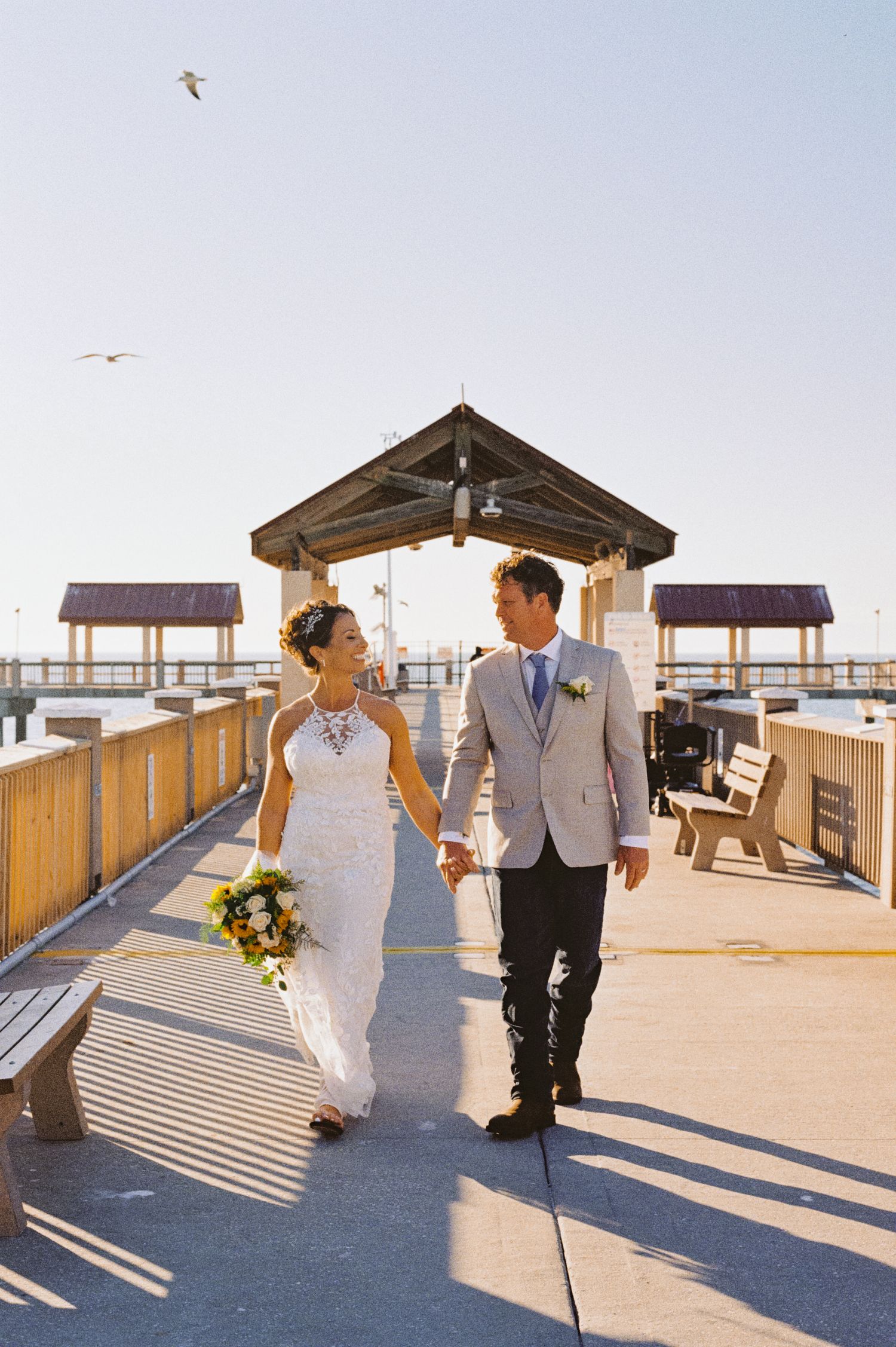 Bride and groom walking hand in hand on sunny pier with seagulls overhead.