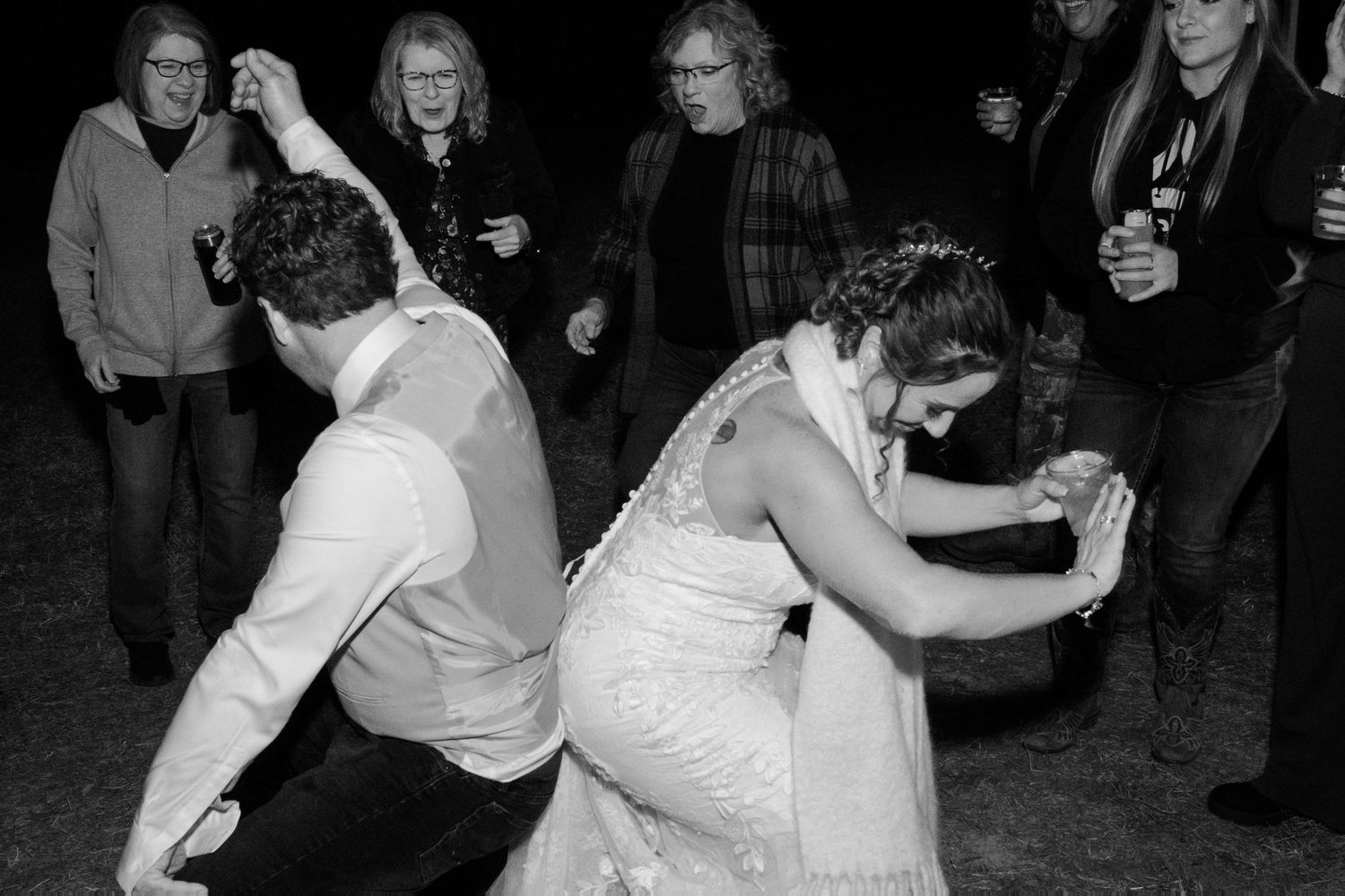 Black and white photo of bride and groom doing limbo dance surrounded by amused wedding guests.