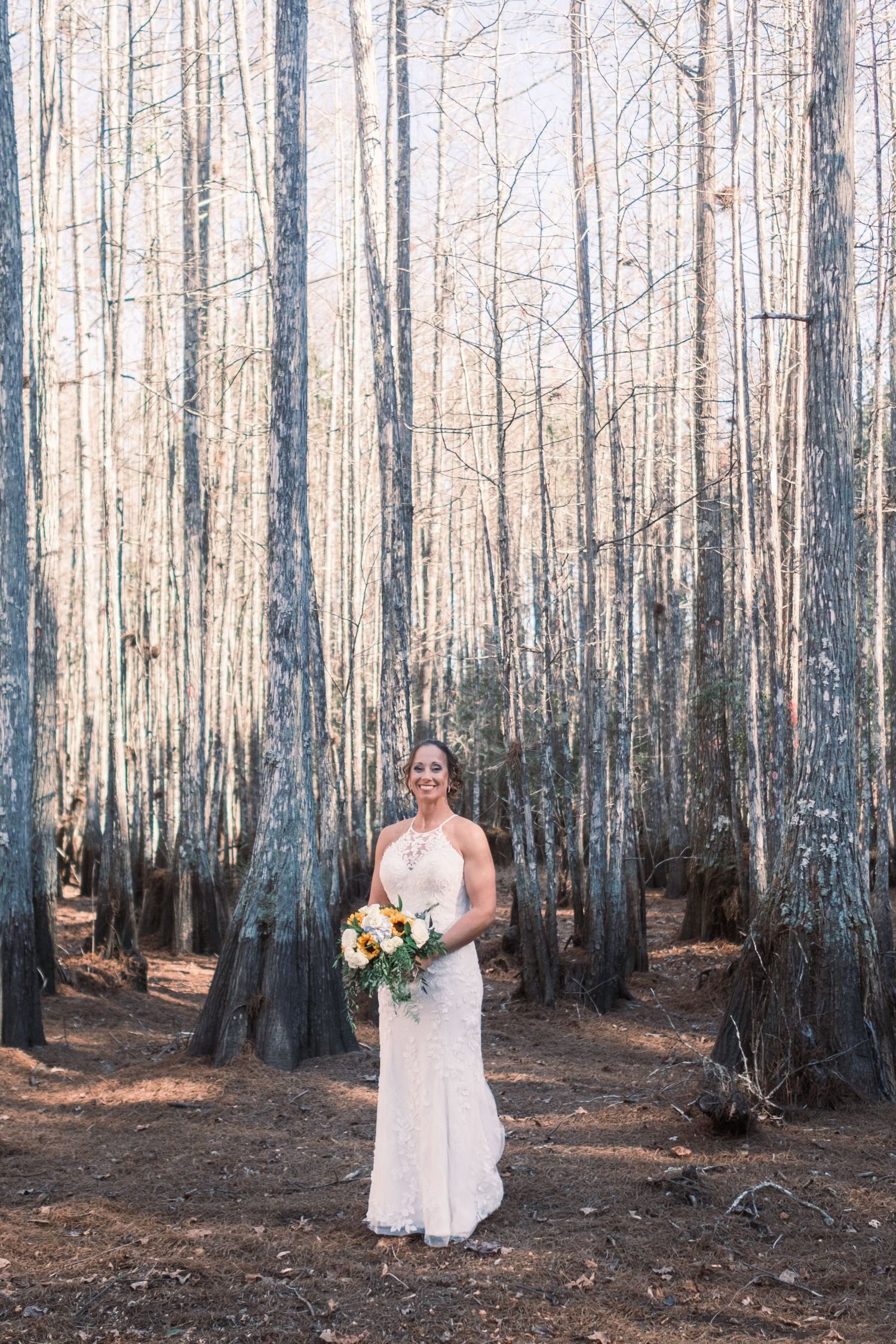 Smiling bride in white lace halter gown holds colorful bouquet while standing among tall bare trees in a forest.