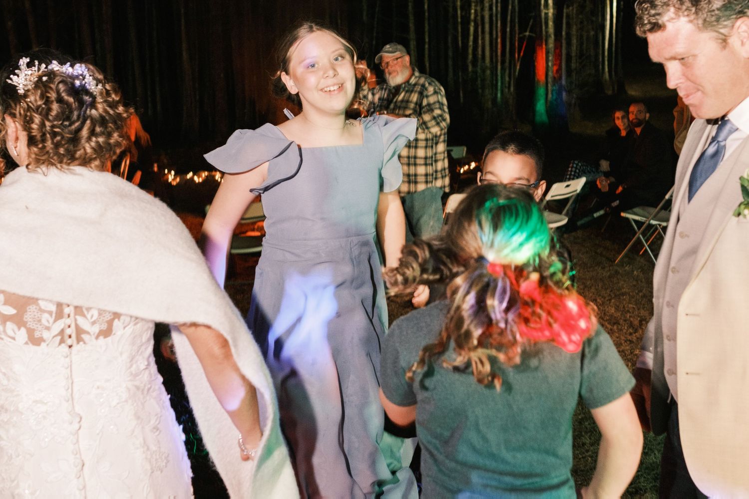Young girl in blue dress dancing happily with wedding guests at an outdoor evening wedding celebration.
