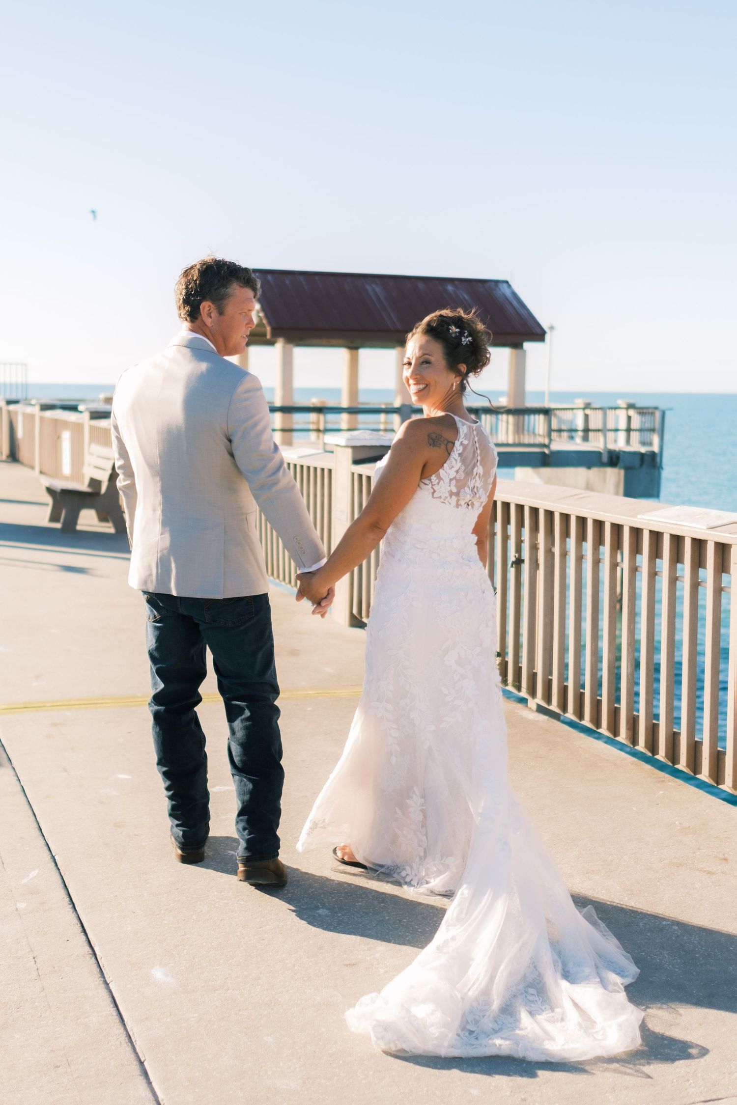 Bride in lace mermaid gown holds groom's hand walking on sunny pier overlooking ocean.