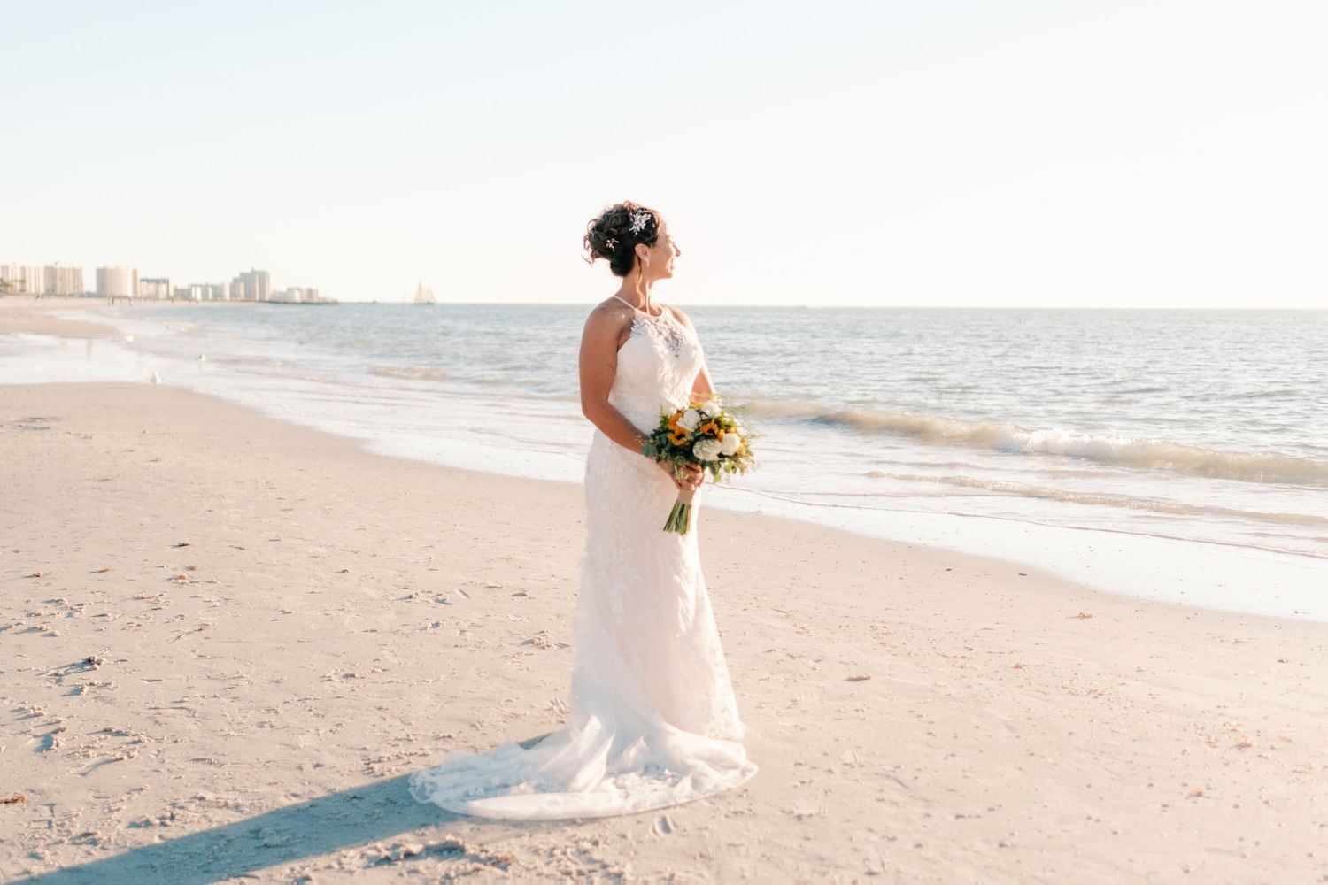 Bride in halter wedding gown holds bouquet while standing alone on a sunny beach.