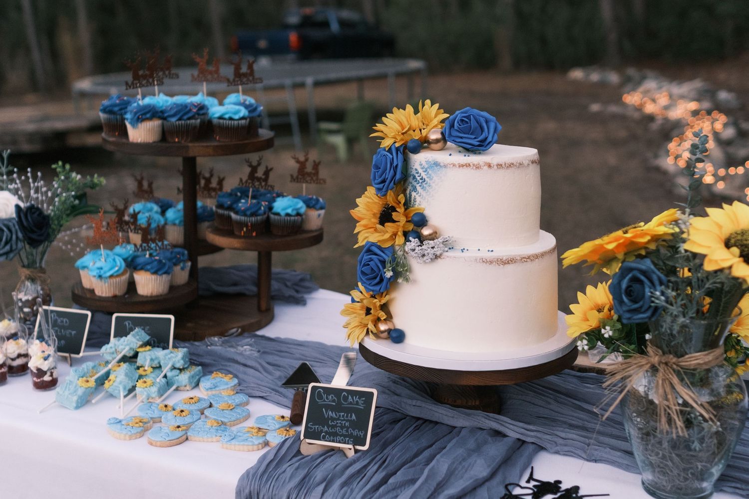 Elegant two-tier white wedding cake decorated with blue roses and sunflowers beside matching blue frosted cupcakes.