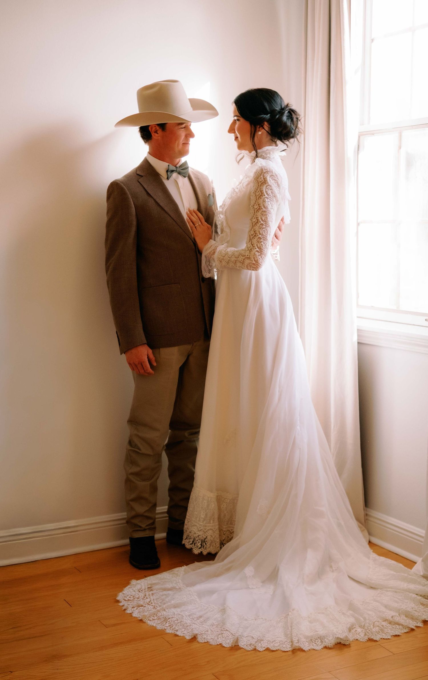 Bride in lace wedding gown with train and groom in tan suit and cowboy hat share a romantic moment by a window.