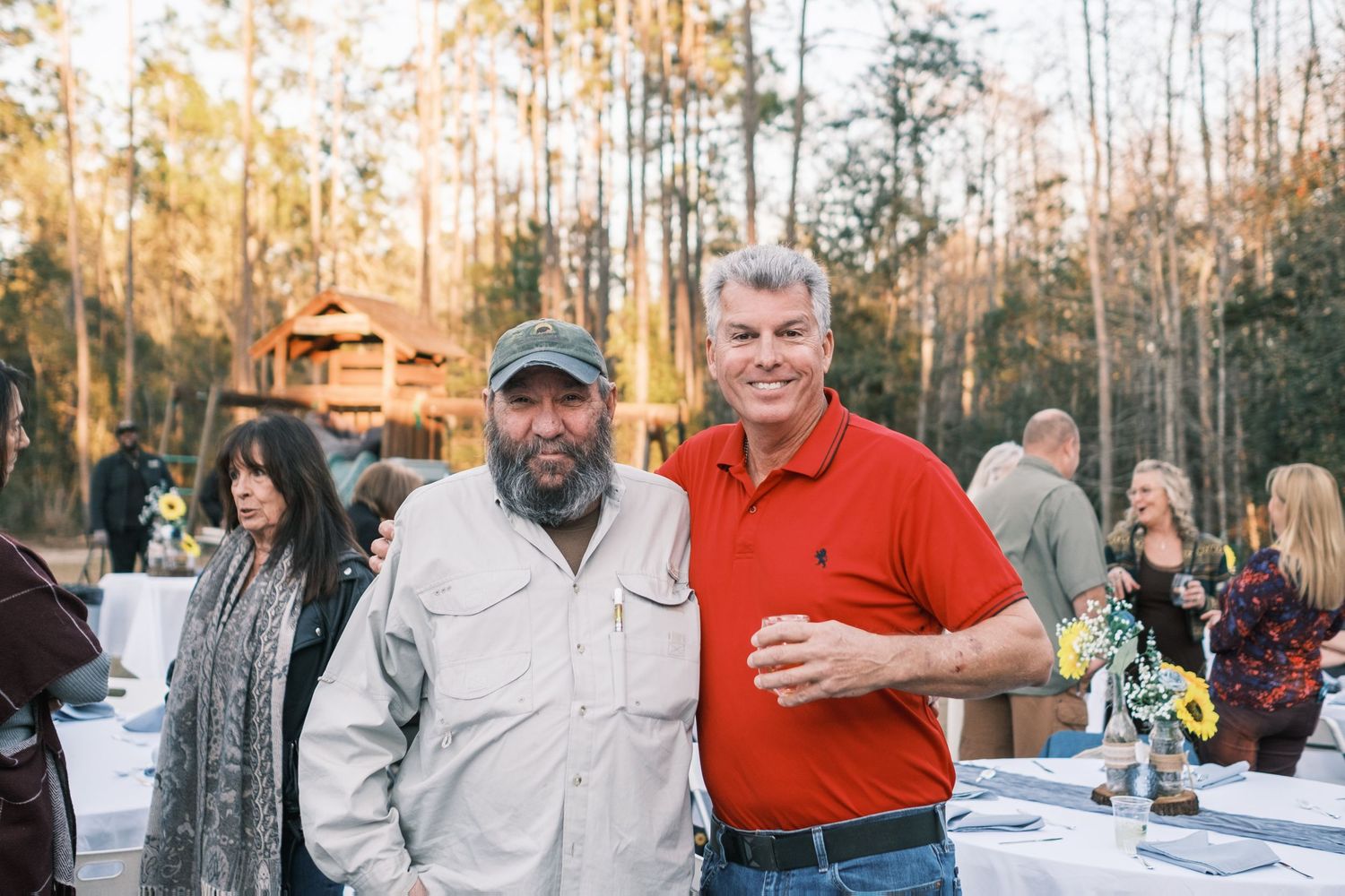Two men smiling and posing together at an outdoor wedding reception surrounded by guests and trees.
