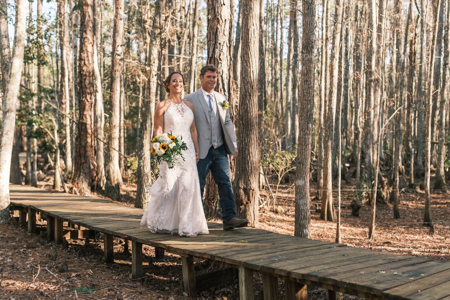 Bride and groom walk hand in hand along wooden boardwalk through sunlit pine forest.