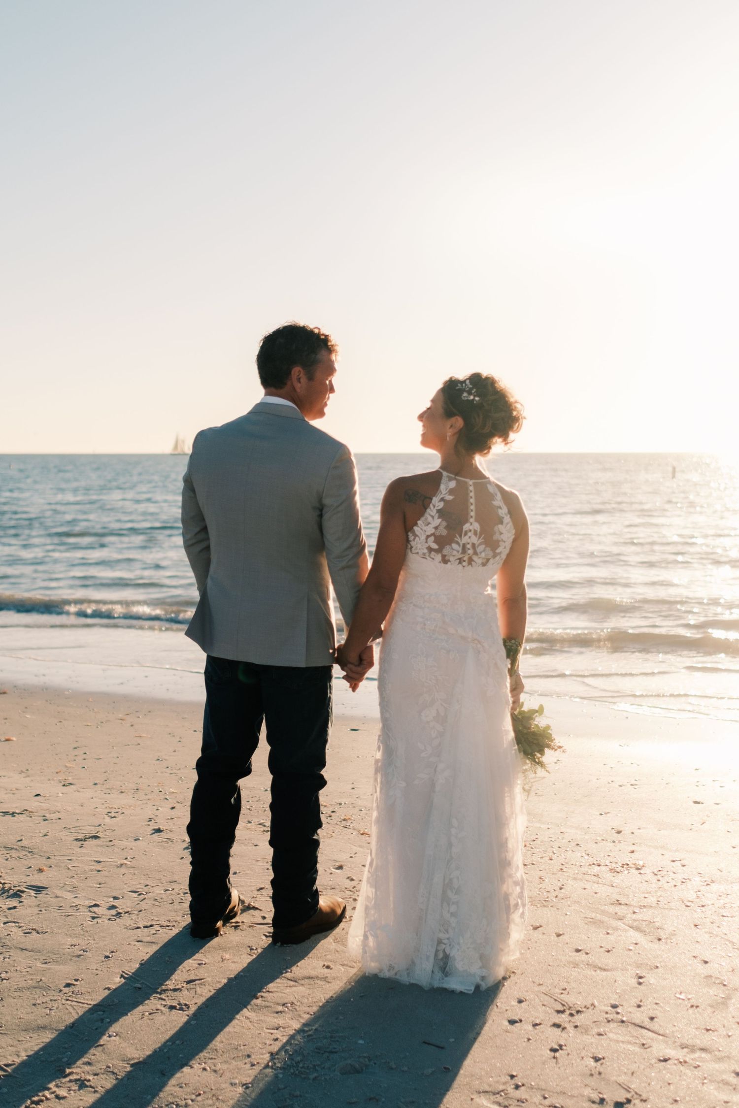 Bride and groom hold hands facing the ocean at sunset on a sandy beach, bride in lace halter gown.