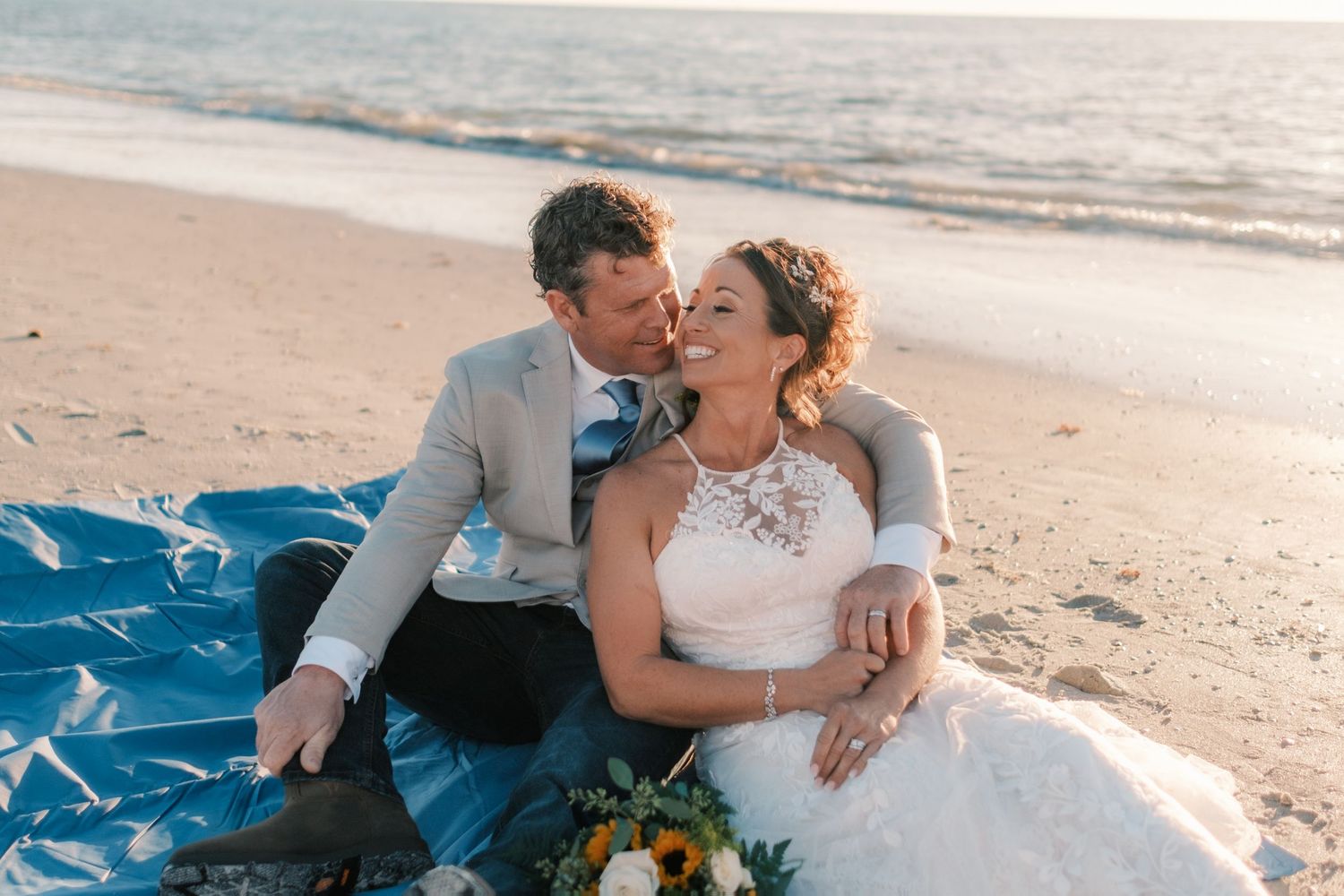 Couple sits on beach blanket at golden hour, groom nuzzling smiling bride holding sunflower bouquet.