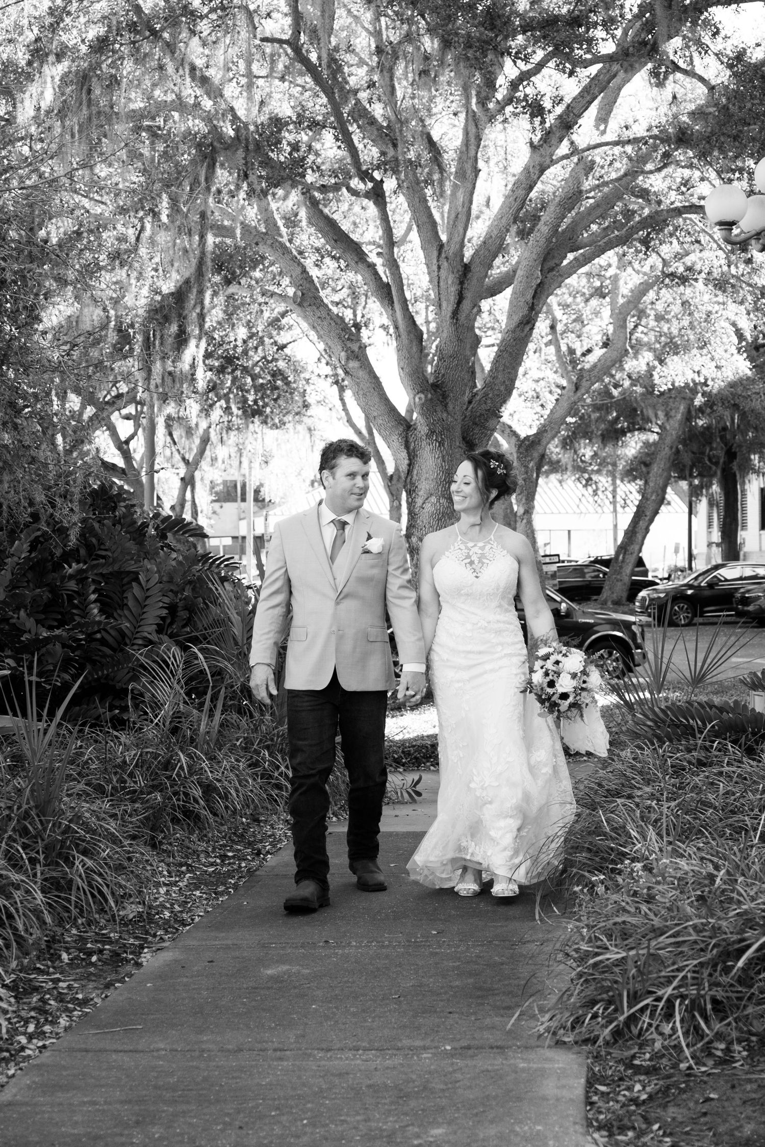 Black and white photo of bride and groom holding hands walking on a path under large oak trees.