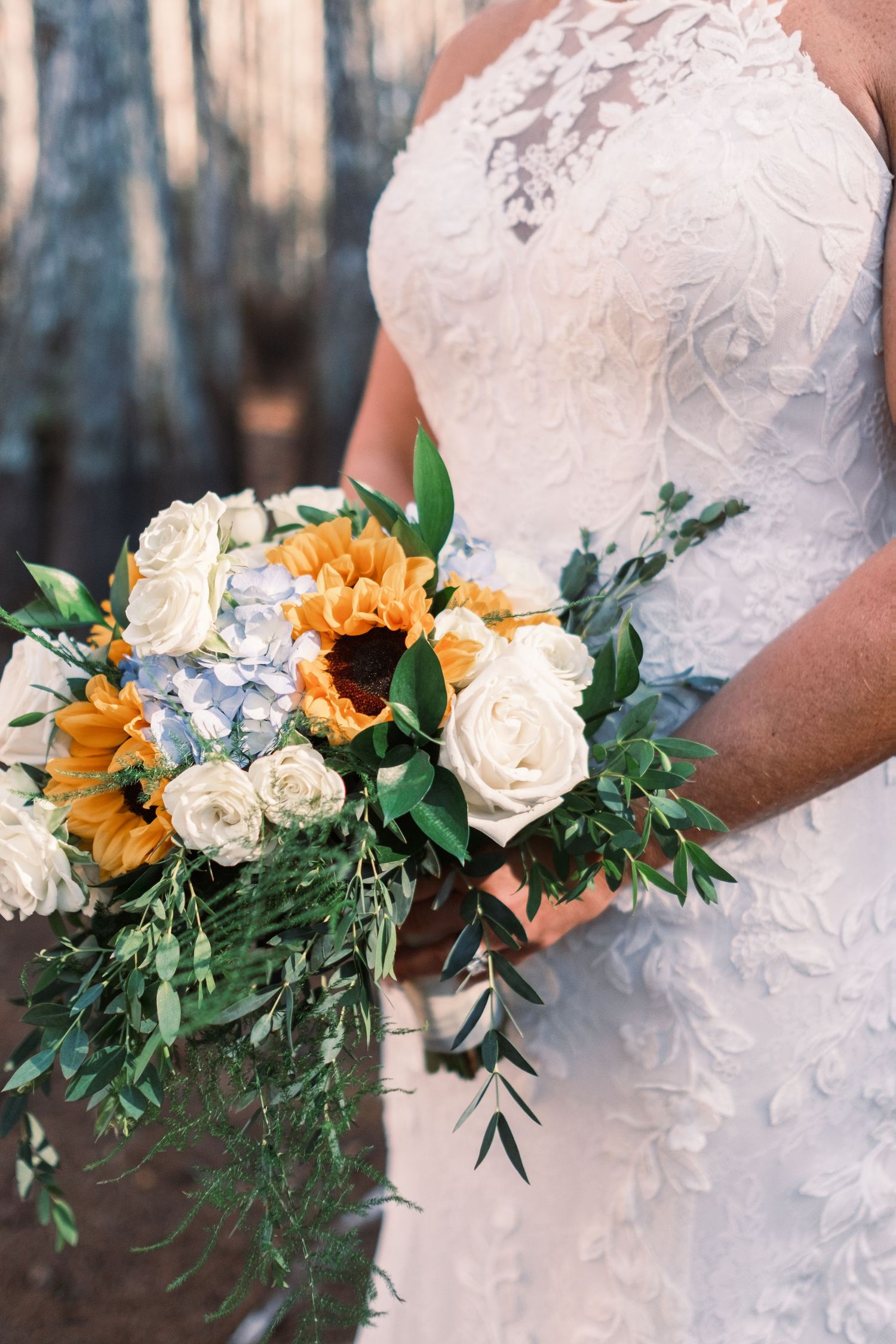 Bride holding a stunning wedding bouquet with sunflowers, white roses, blue hydrangeas, and greenery in a lace gown.