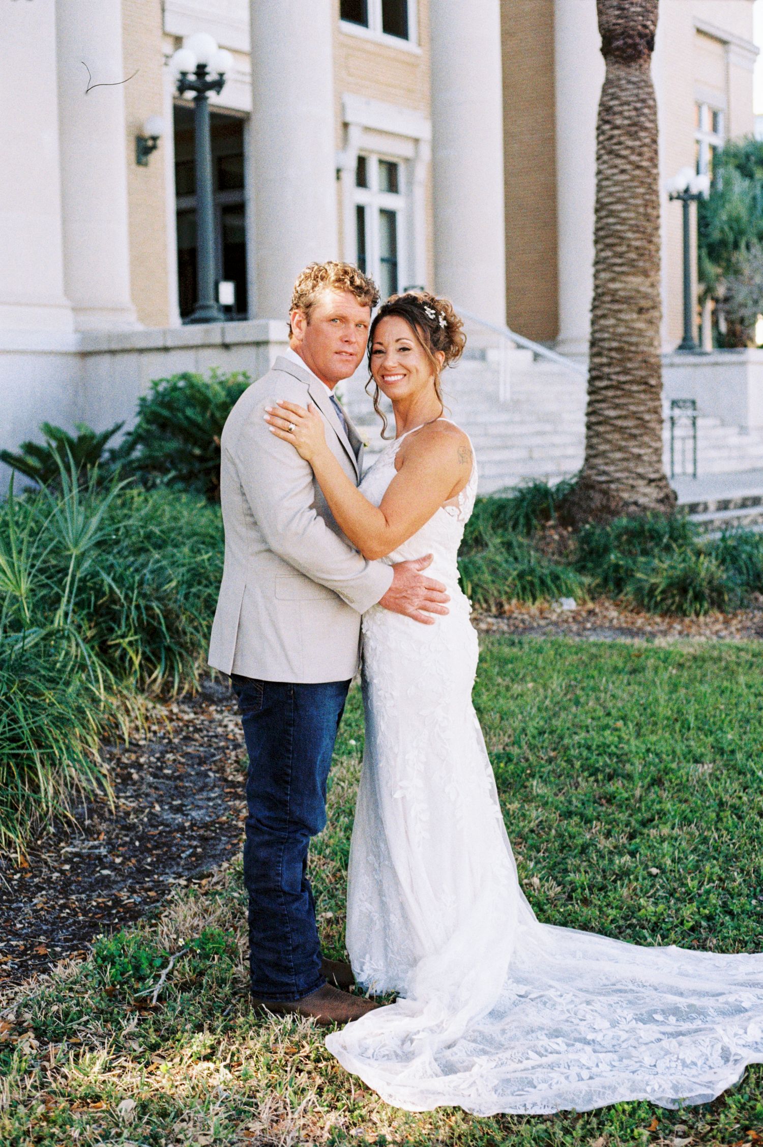 Smiling bride and groom embrace outdoors in front of elegant building with palm tree on wedding day.