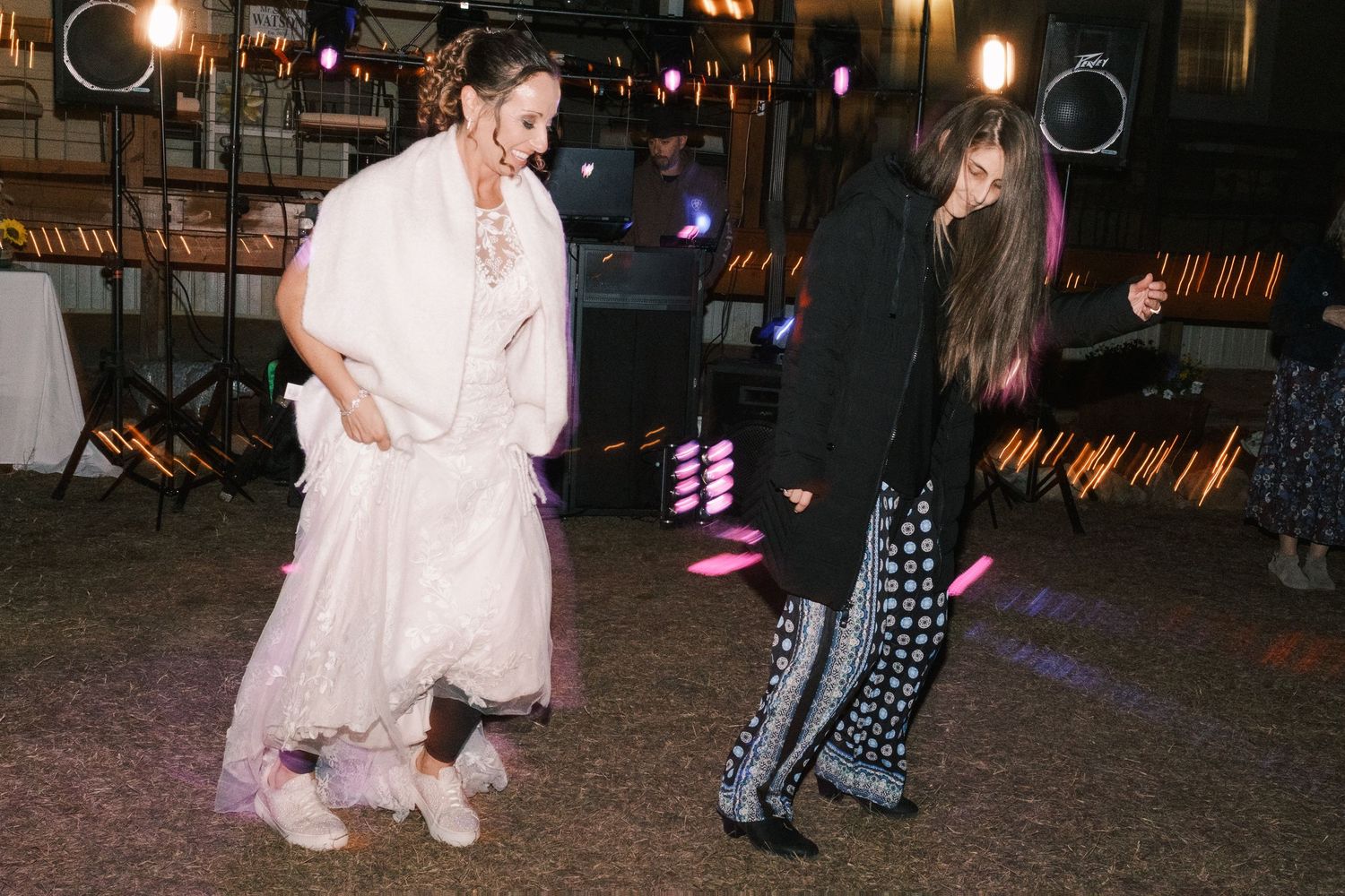 Bride in white gown dances energetically with a guest under colorful lights at wedding reception.
