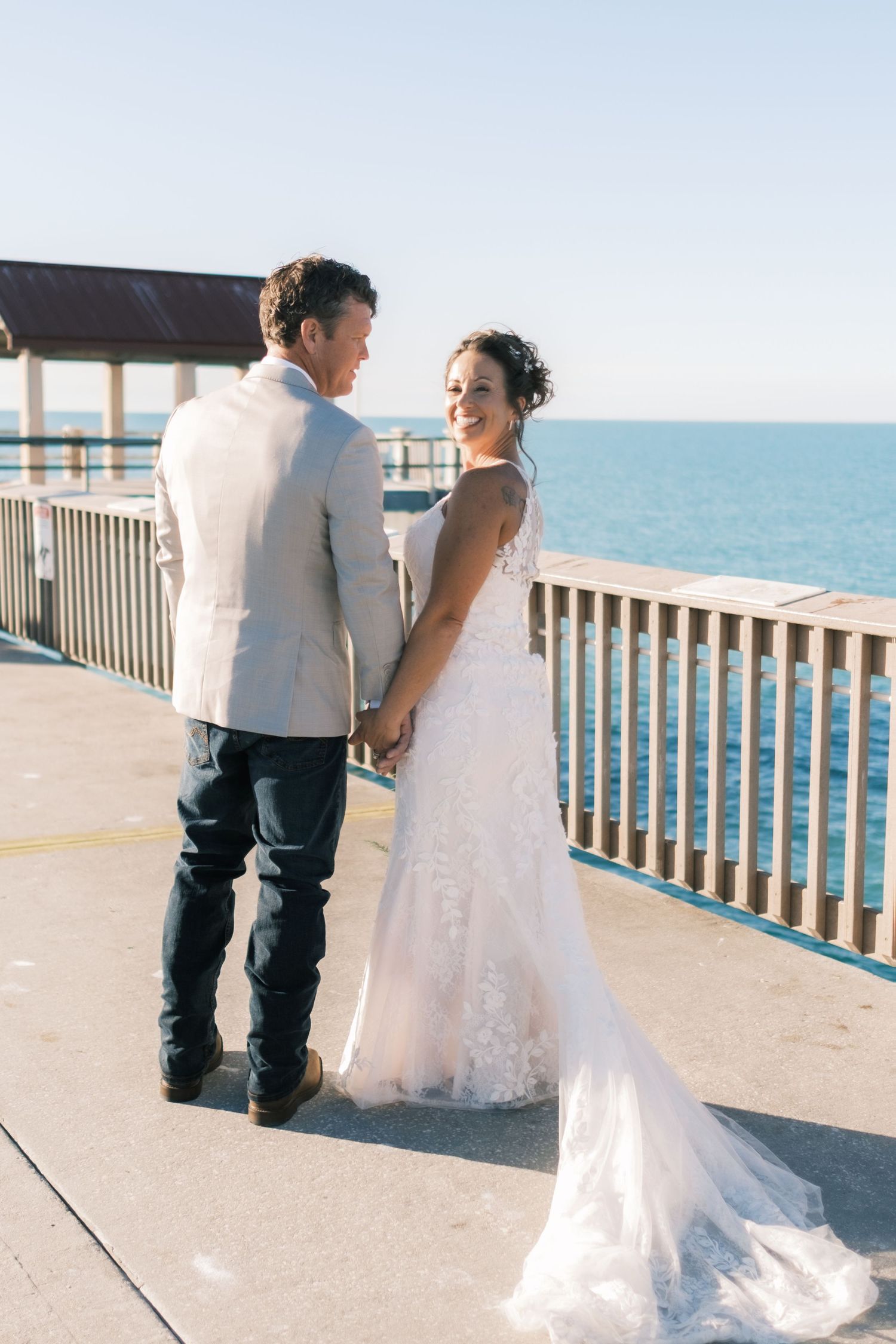 Bride and groom hold hands on a sunny pier overlooking the ocean during wedding portraits.