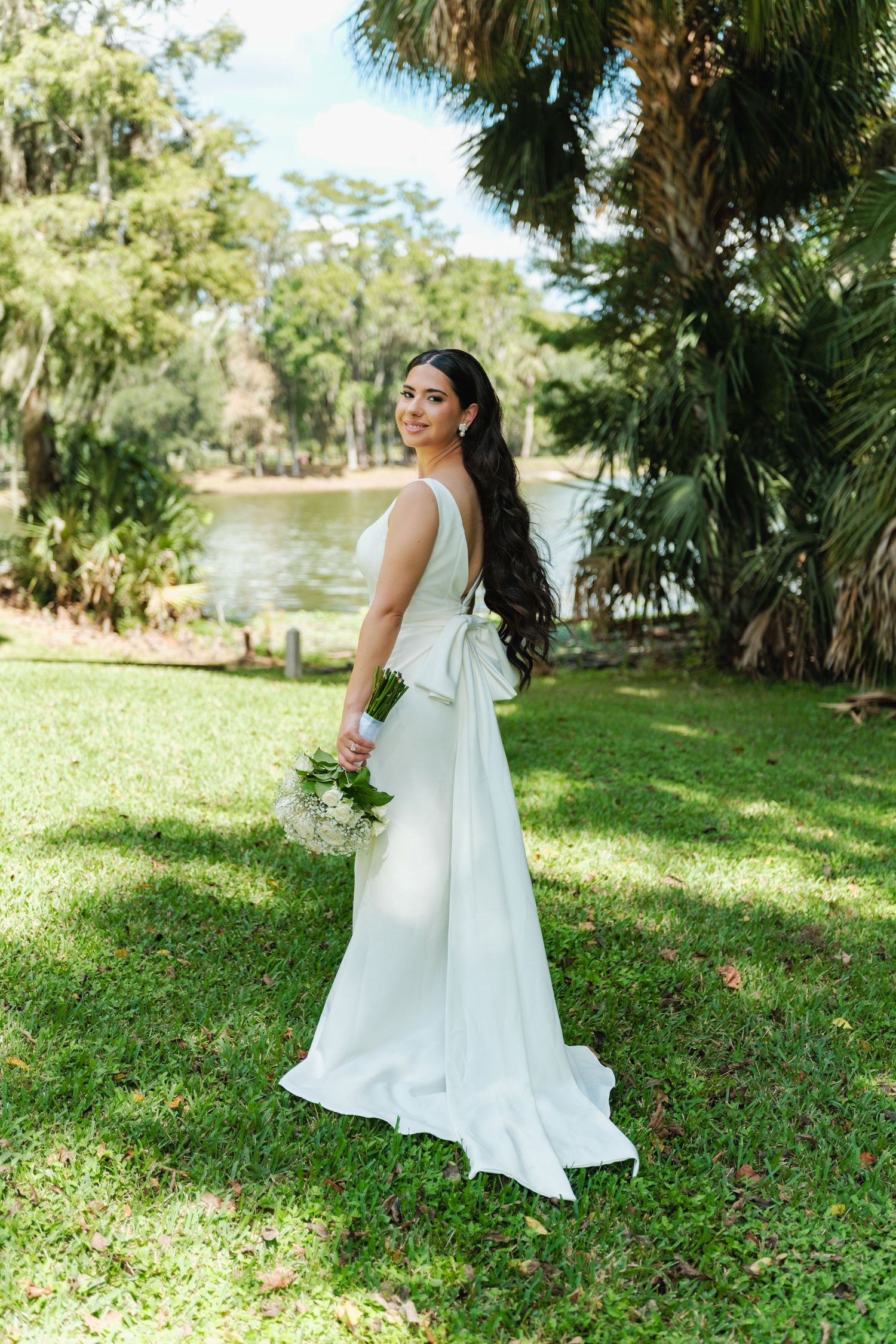 A flowing white wedding dress moves gracefully across a sunlit lawn near a lake.