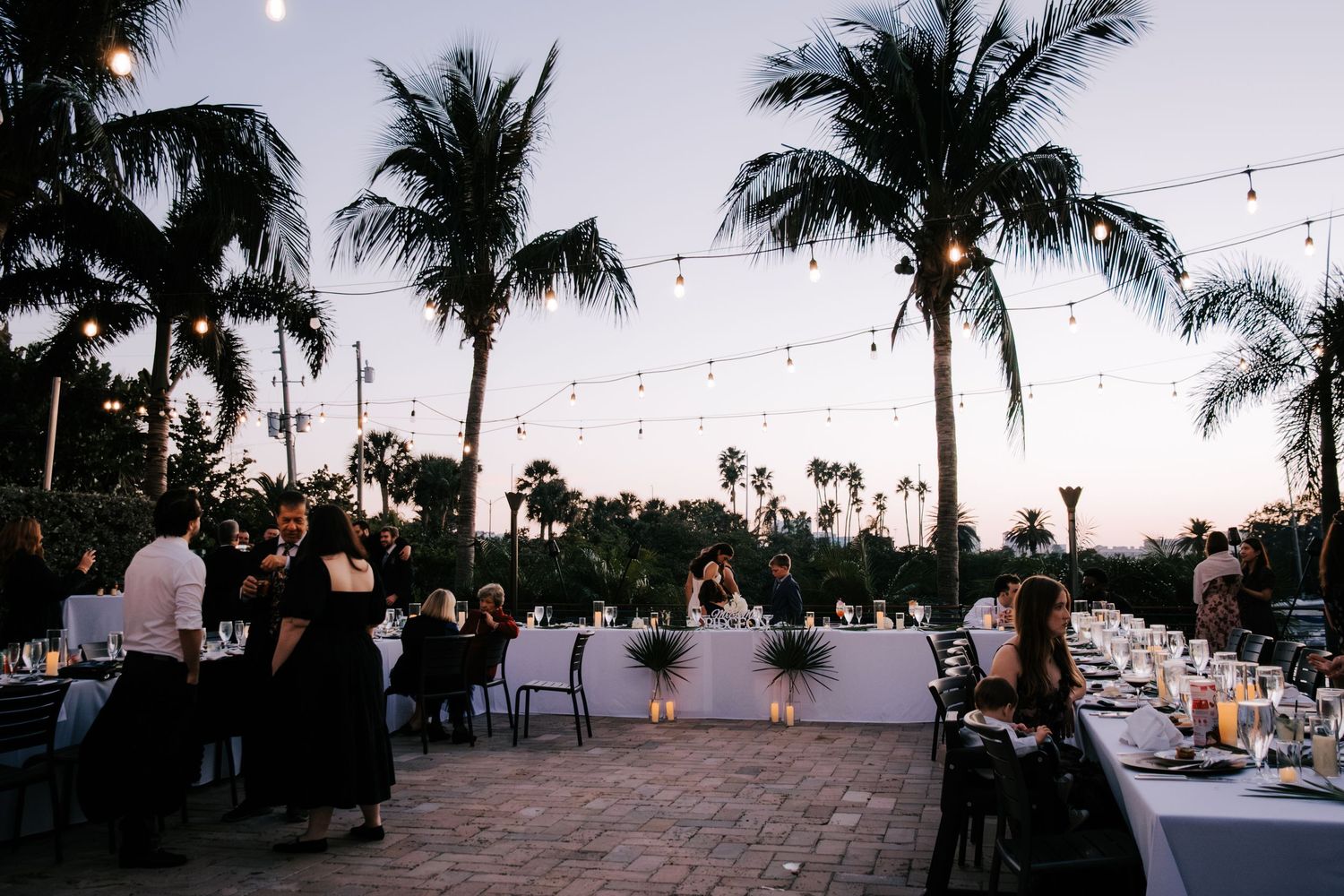 Palm trees silhouetted against sunset sky with string lights at outdoor wedding reception.