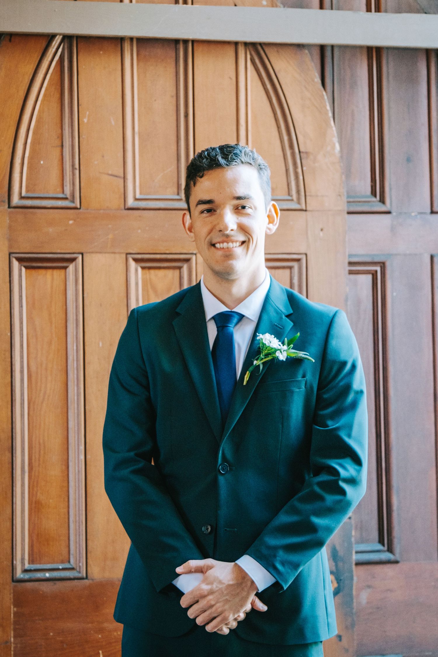 Groomsmen in matching dark suits stand in a row against a wood-paneled wall at a formal wedding.