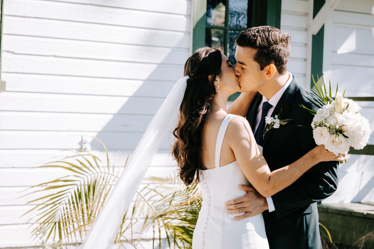 Wedding ceremony moment captured outside a white church building with palm fronds in the background.