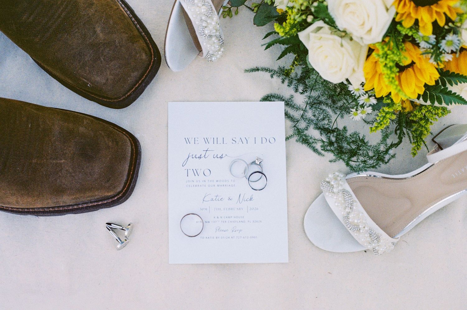 Wedding flat lay with cowboy boots, heels, sunflower bouquet, rings, and save the date card.