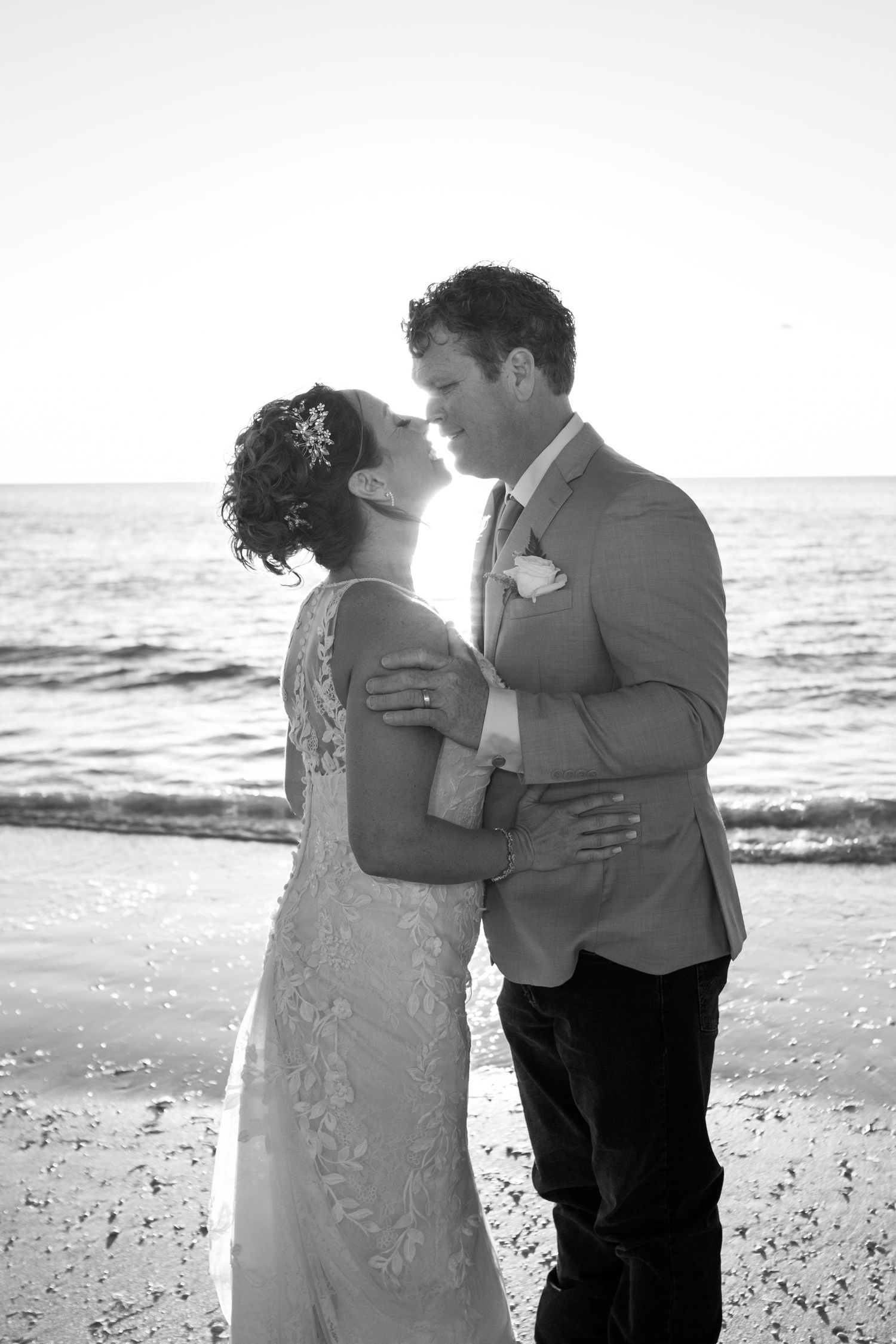 Black and white photo of bride and groom sharing a tender kiss on the beach at sunset.