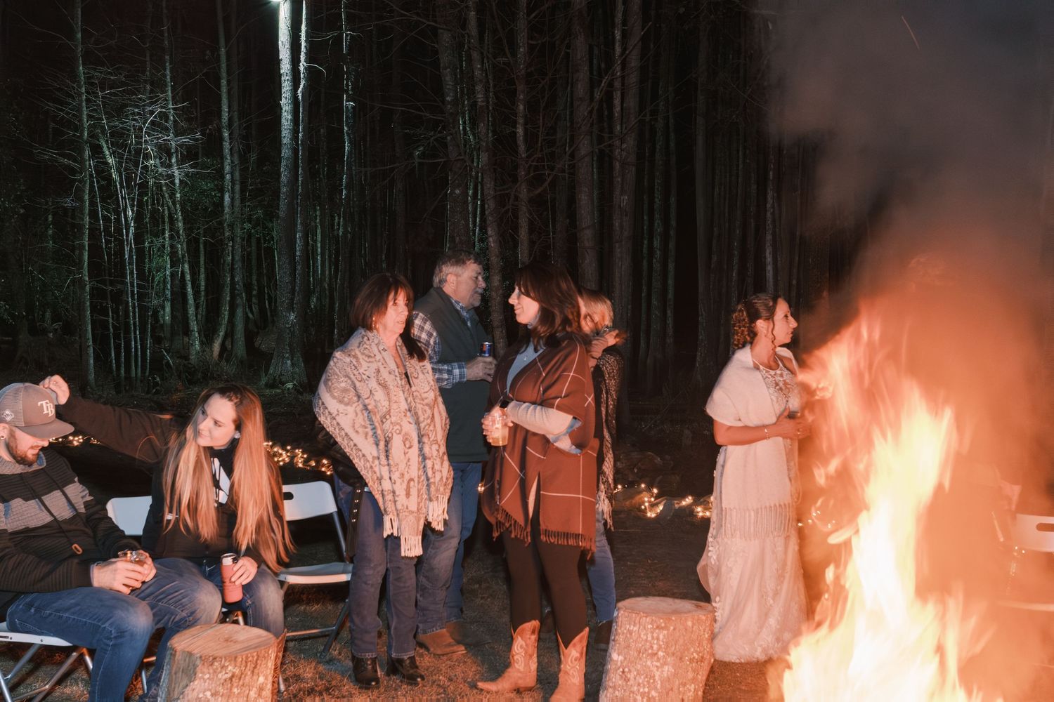 Wedding guests gather around a large bonfire at night in a forest during an outdoor reception celebration.
