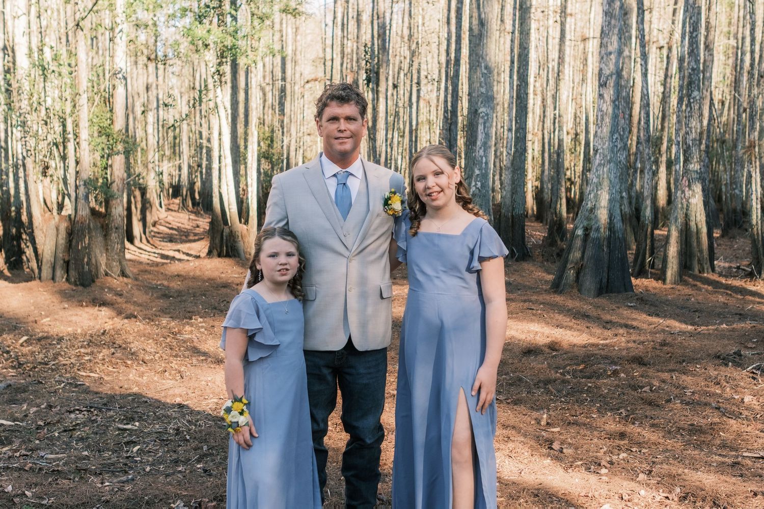 Groom in tan suit poses with two young girls in blue dresses in a wooded cypress forest.