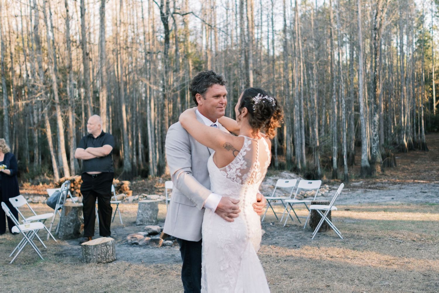 Bride and groom share first dance outdoors at twilight with white chairs and fire pit in background.